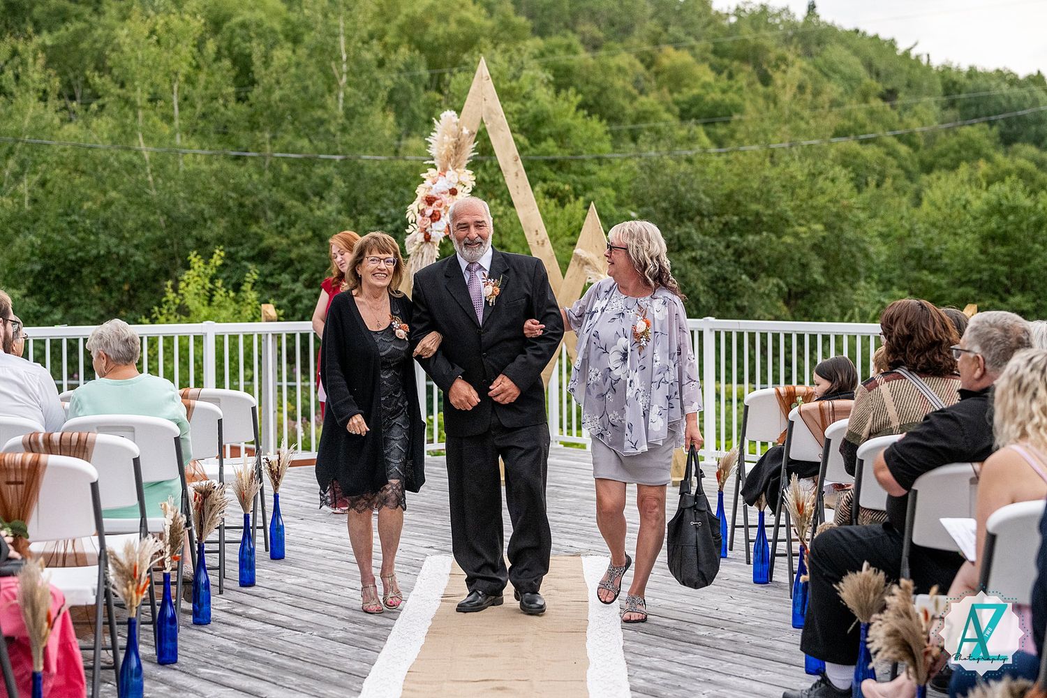 Wedding ceremony guests gathered on a white deck with blue decorative accents and green foliage backdrop.
