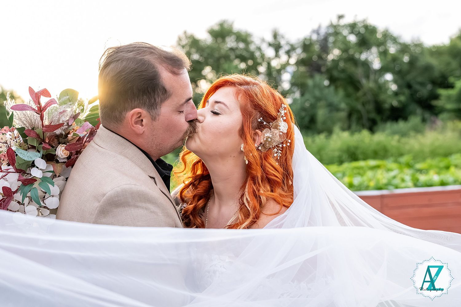 Romantic couple shares intimate kiss under bridal veil on outdoor deck with sunset lighting.