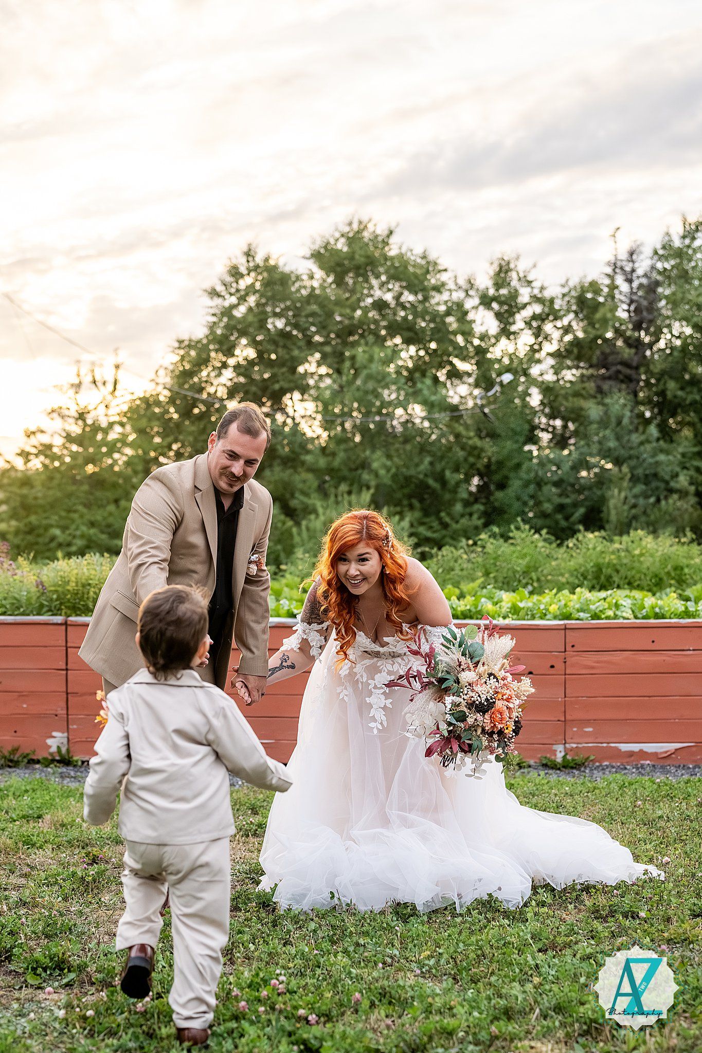 Couple in wedding attire share playful moments together with their young child on grass during sunset photo session.