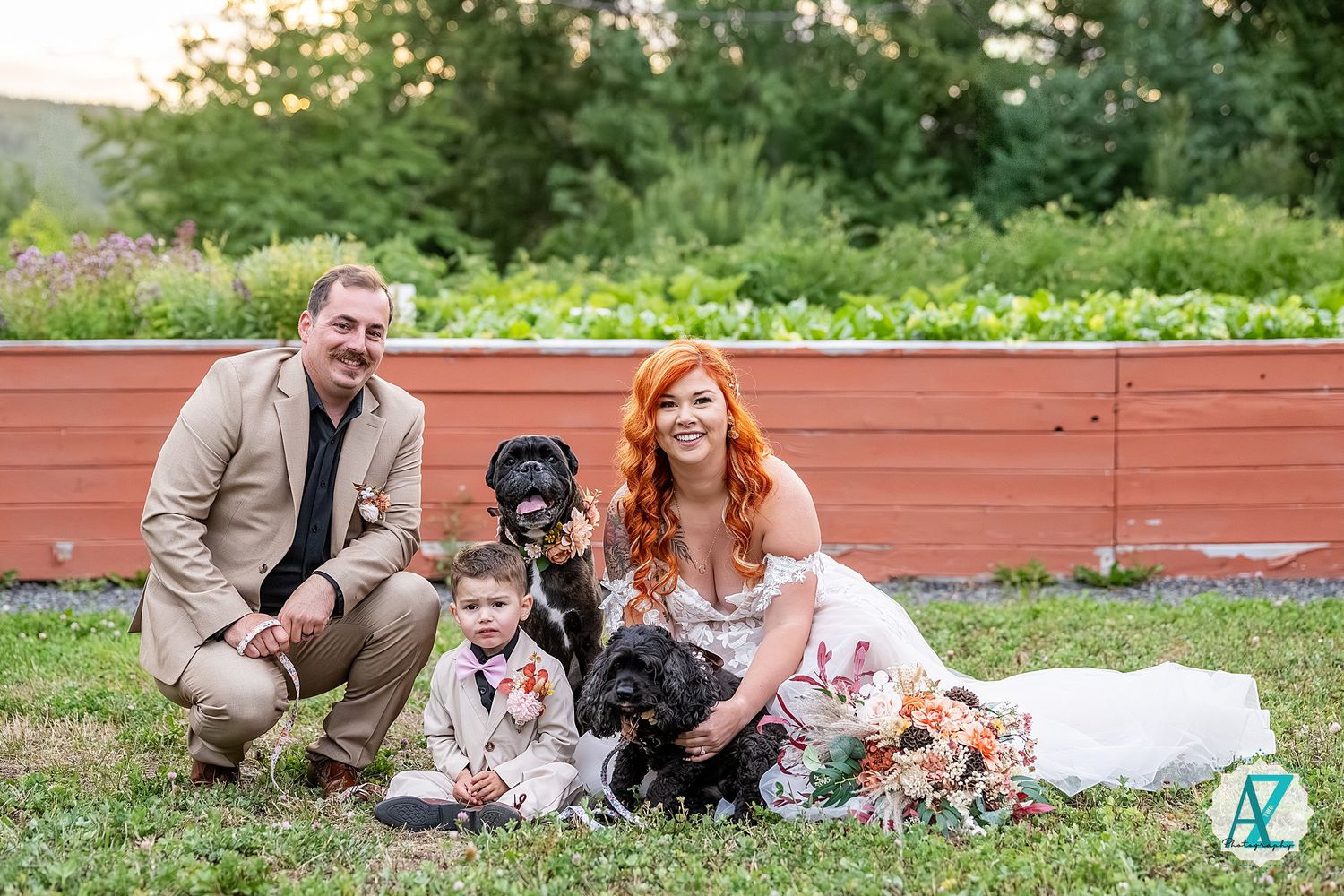 A couple and their dog pose together on grass near red wooden fencing during an outdoor photo session.