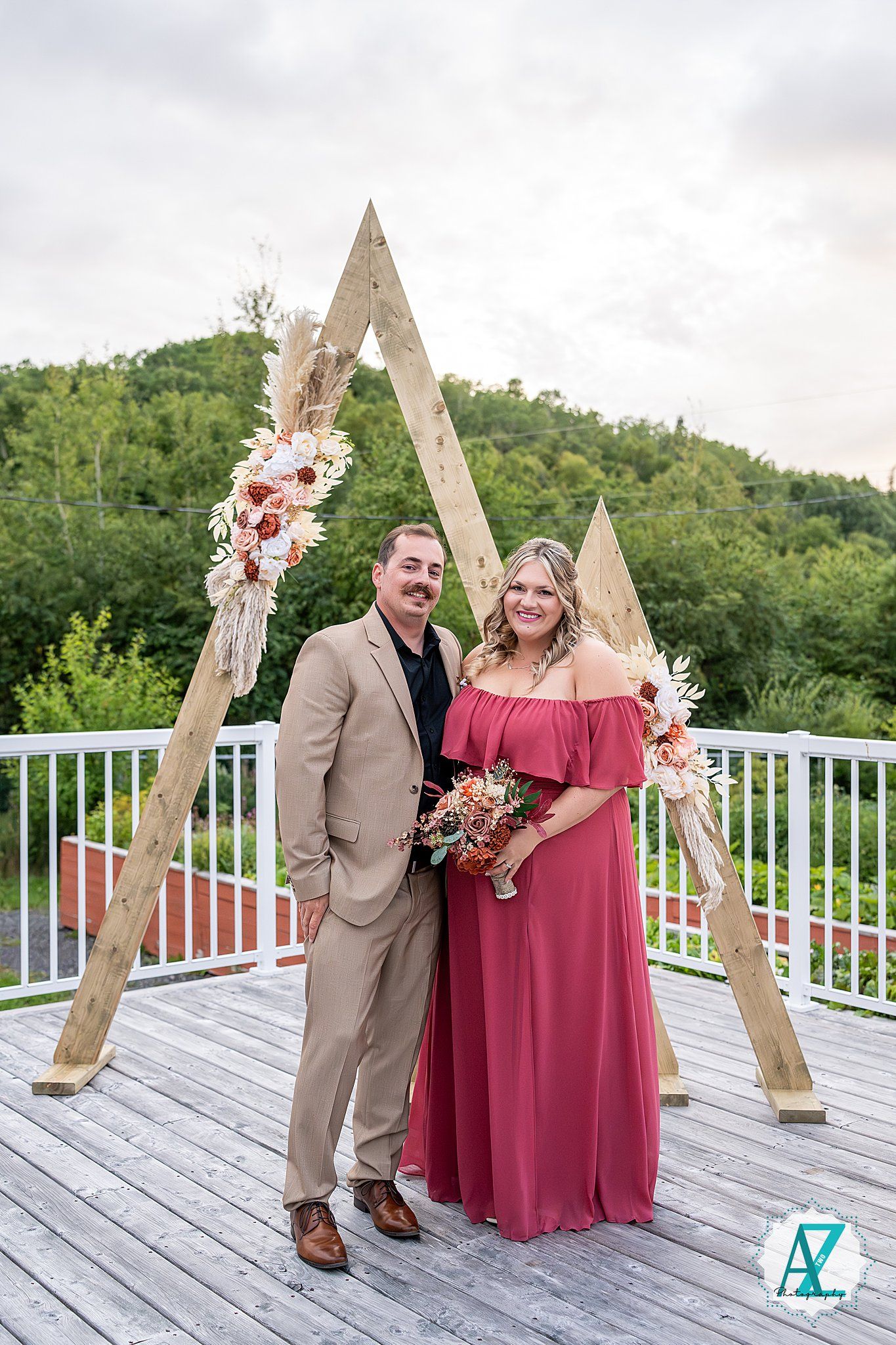 Couple poses by wooden triangle arch on deck with pink and burgundy wedding decor.