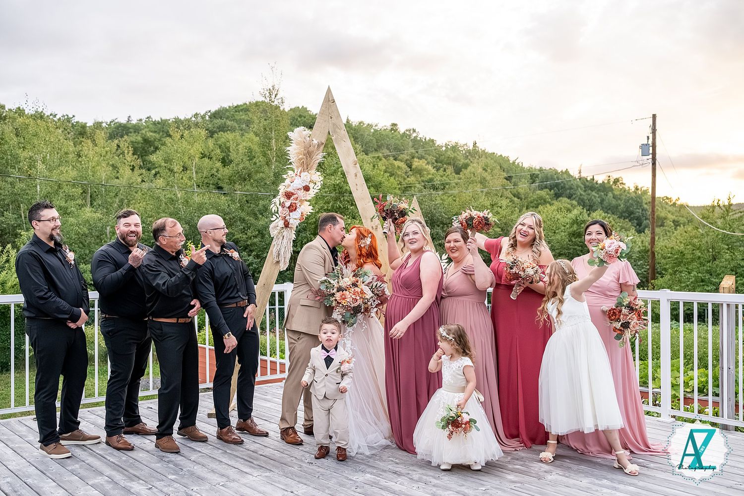 Wedding party in pink and black attire pose together on outdoor deck with mountain views at sunset.