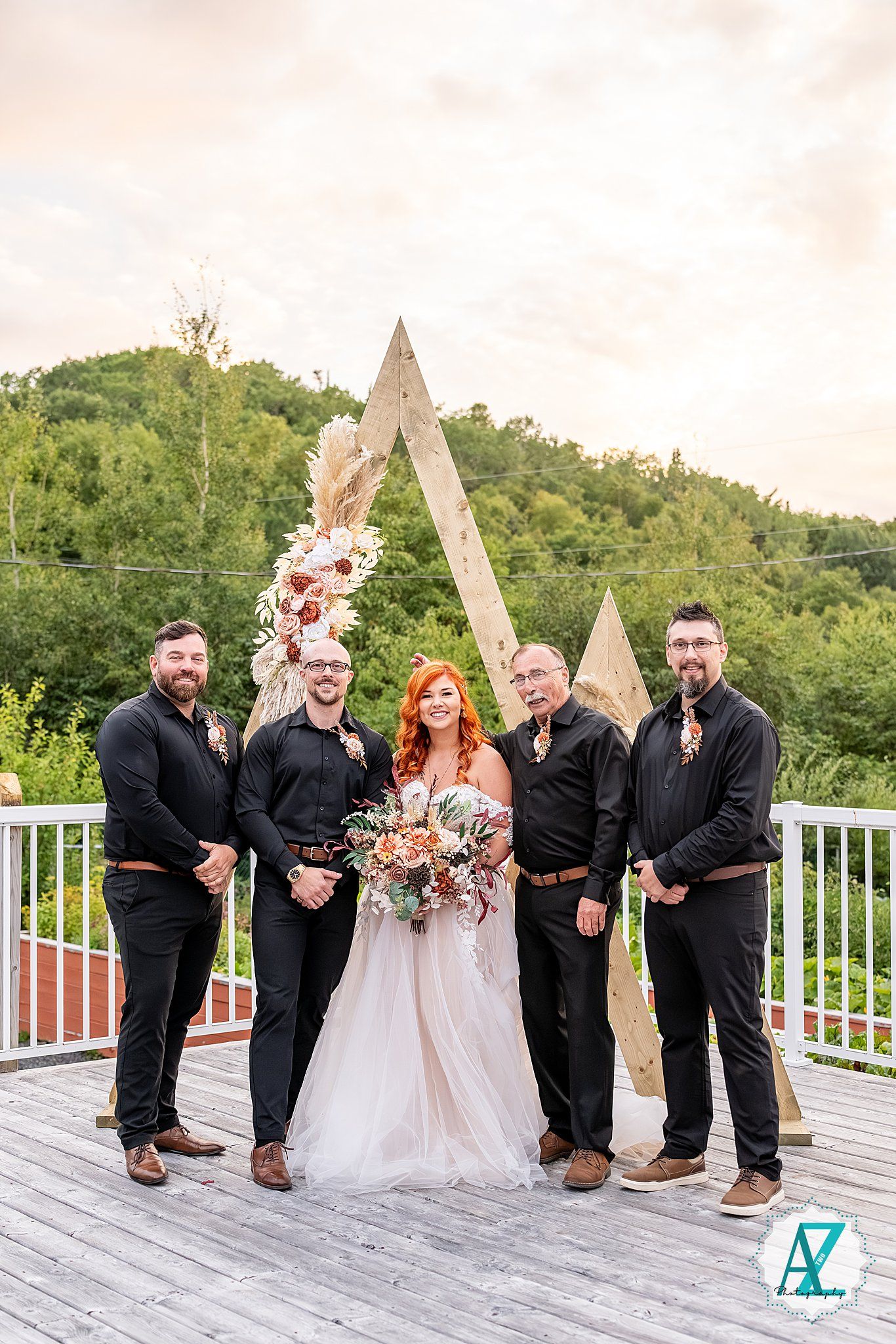 Wedding party in black attire gathers around couple on outdoor deck with wooden arch.