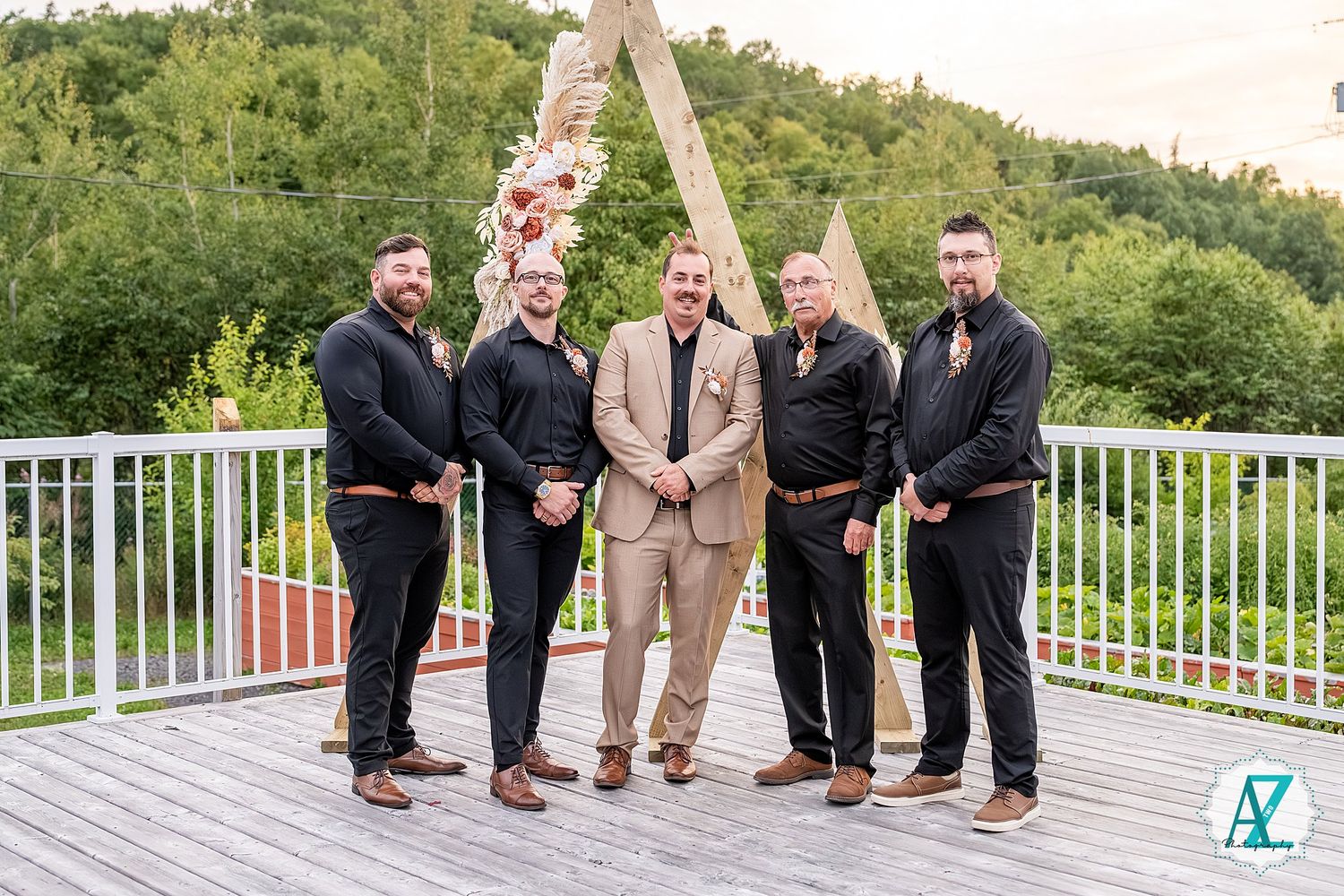 Wedding party in formal attire poses lined up on white deck railing with lush green mountain backdrop.