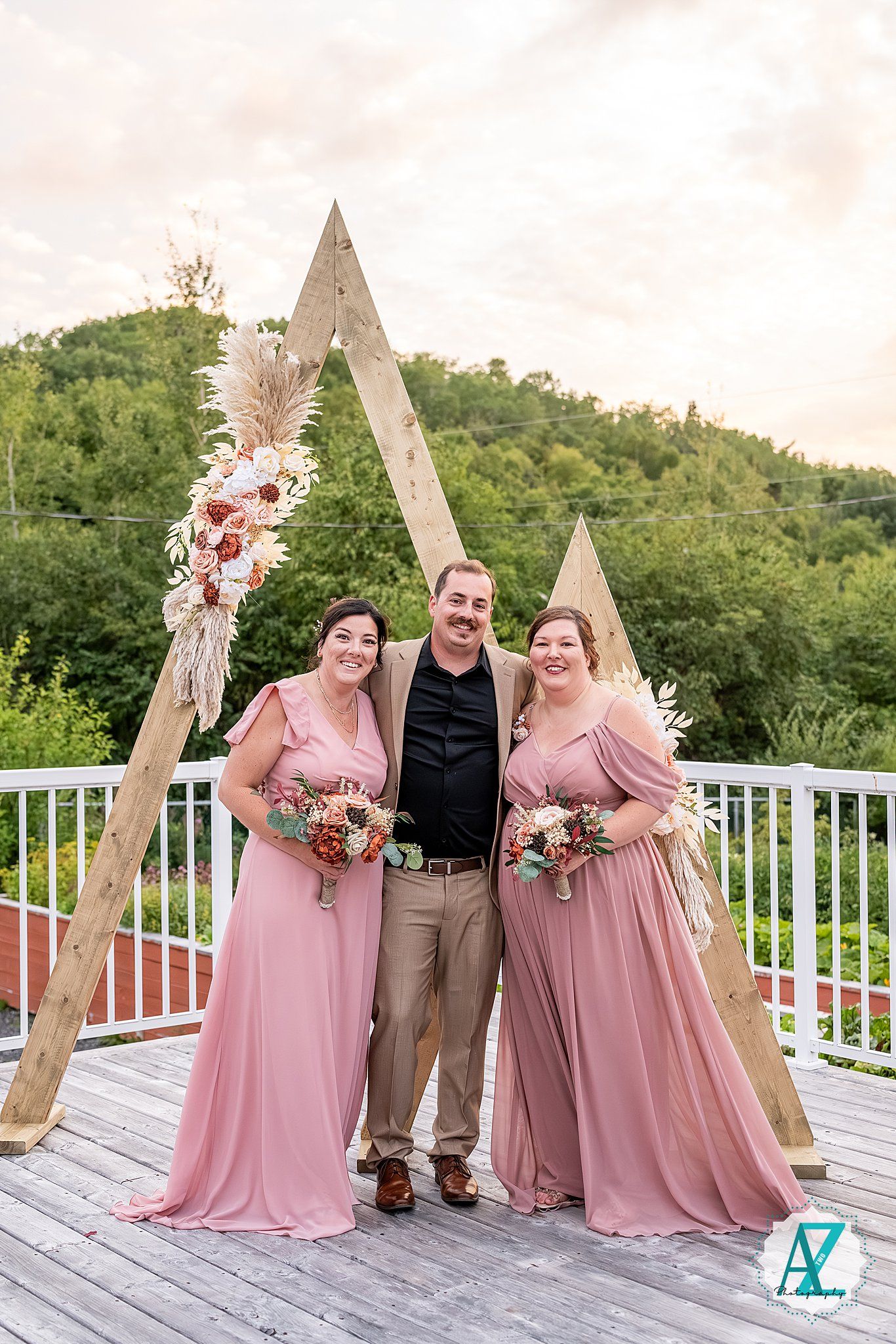 Bridal party in pink dresses poses with wooden triangle arches on outdoor deck.