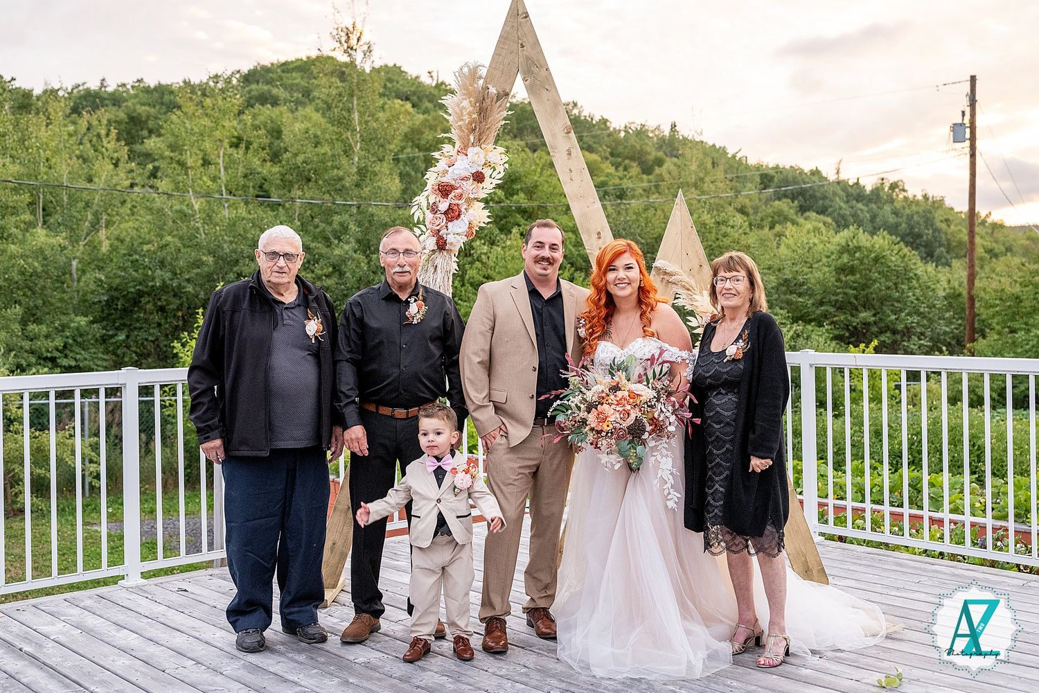 A group of people stand together on a wooden deck with mountainous greenery and an A-frame structure in the background.