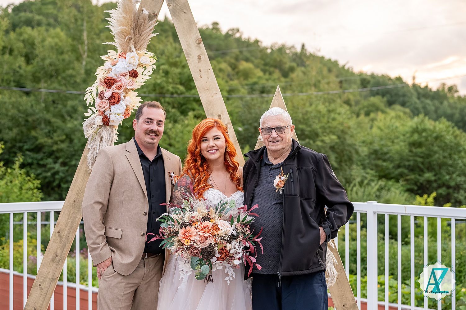 Wedding group photo on outdoor deck with triangle arch and floral decorations.