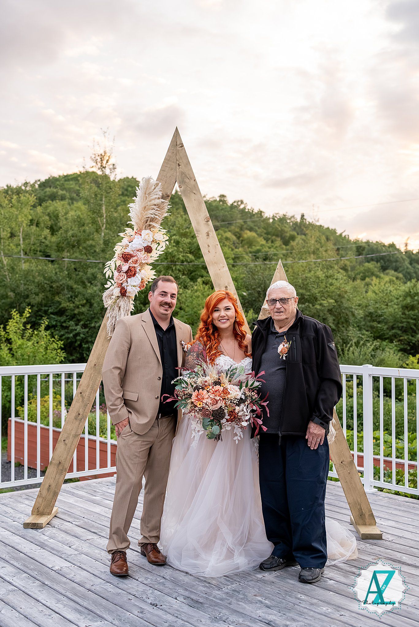 Wedding gathering on wooden deck with triangle arch and lush green backdrop at sunset.