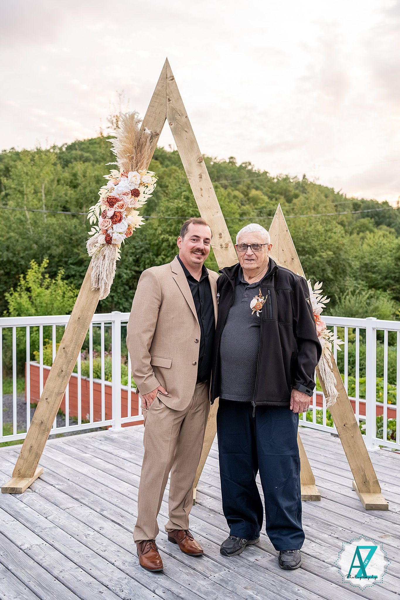 Wedding guests gather near a wooden geometric arch on outdoor white deck with mountain views.