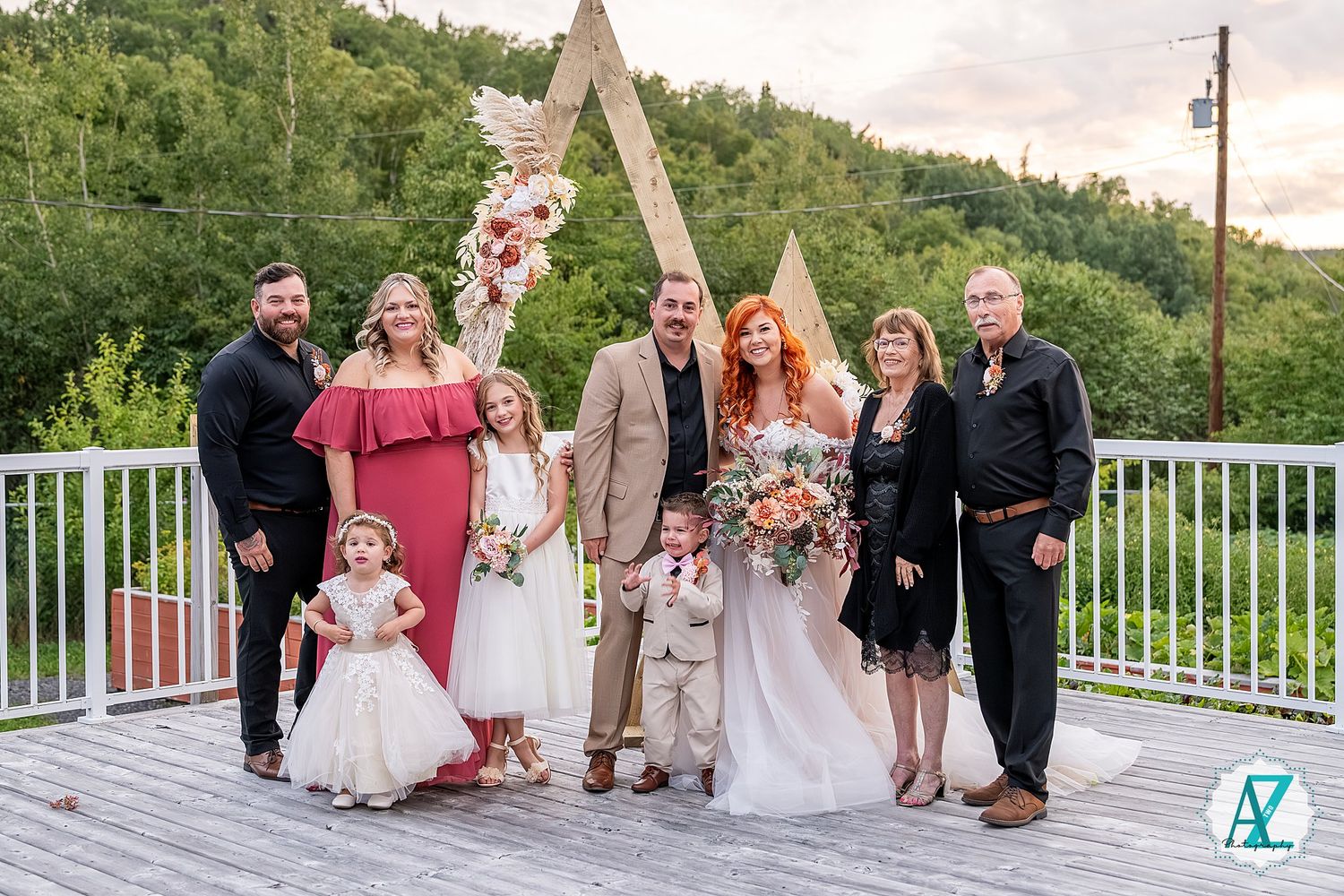 Wedding party gathered on wooden deck with triangular arch decorations and pink bridesmaid dresses.