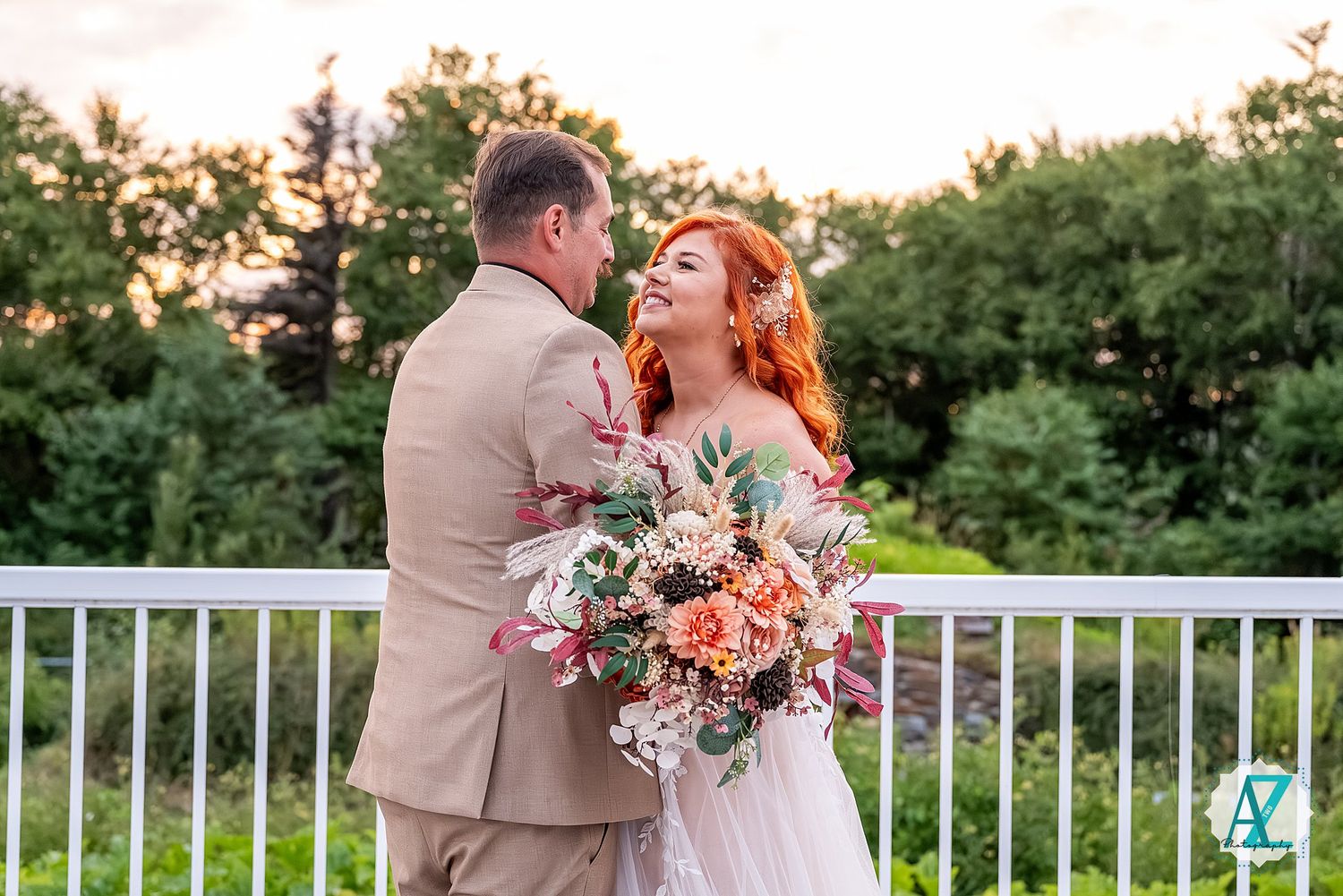Scenic outdoor wedding moment on white deck with floral triangular arch and lush green backdrop.