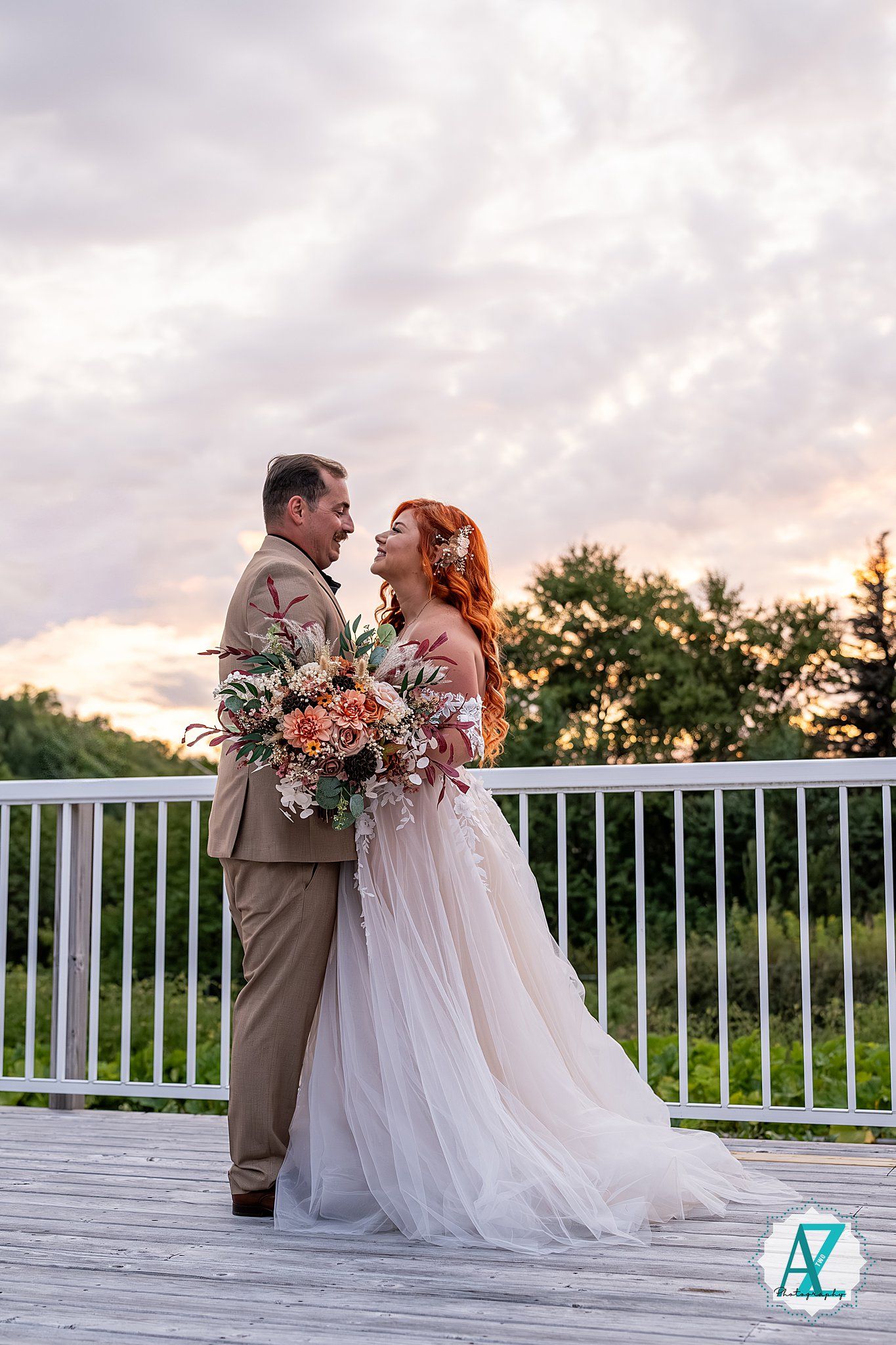 Bride in flowing white gown and groom in tan suit embrace on a deck at sunset holding floral bouquets.