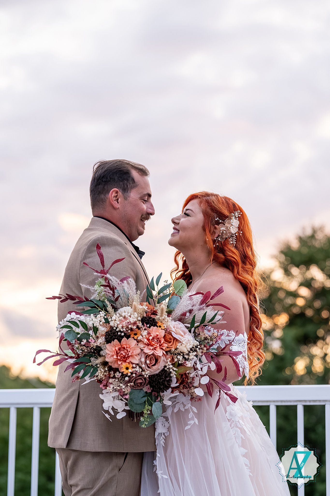 Romantic outdoor wedding scene at sunset showing couple with pink floral bouquet on white deck.
