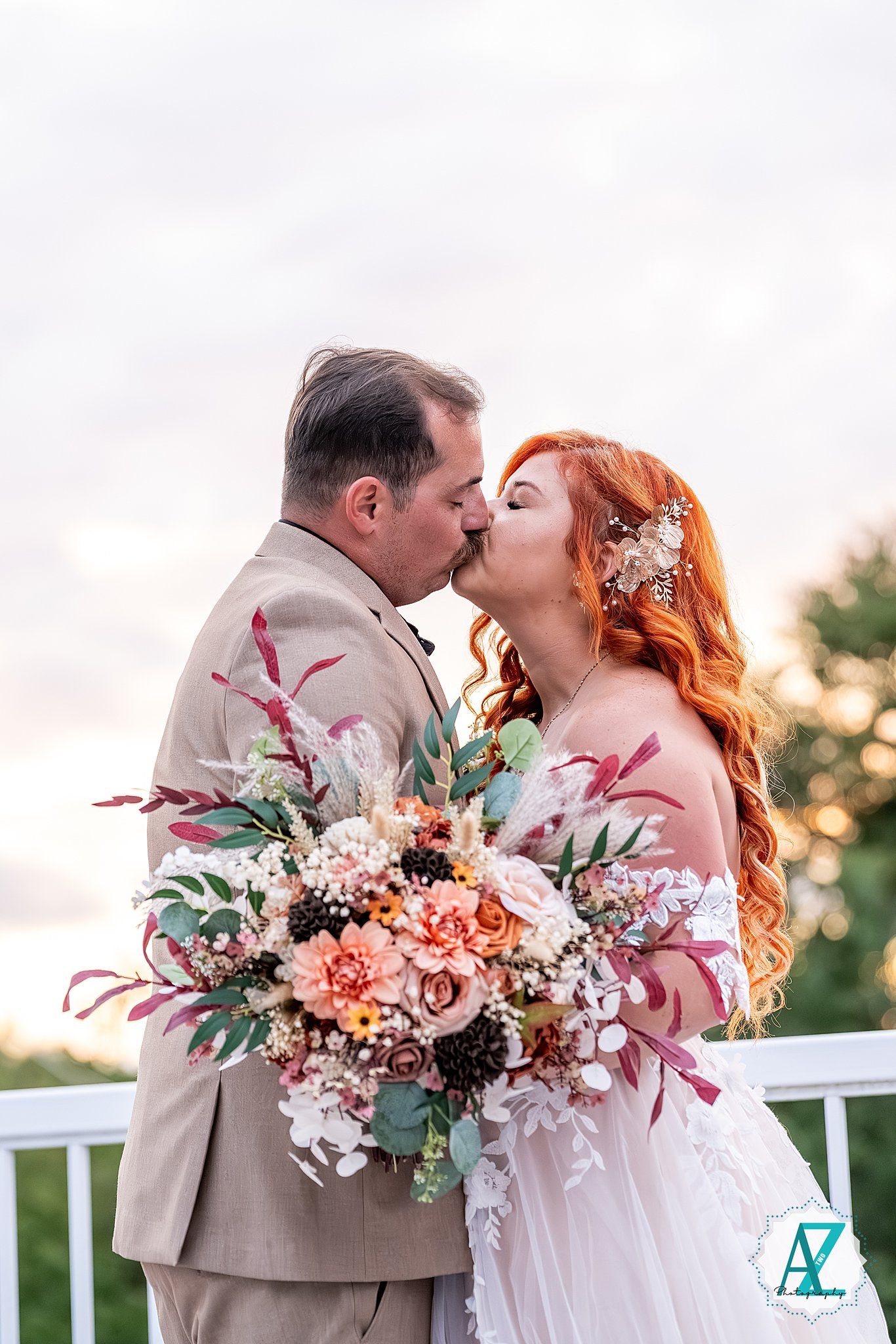 Romantic sunset portrait of newlyweds with pink and peach floral bouquet against white railing.