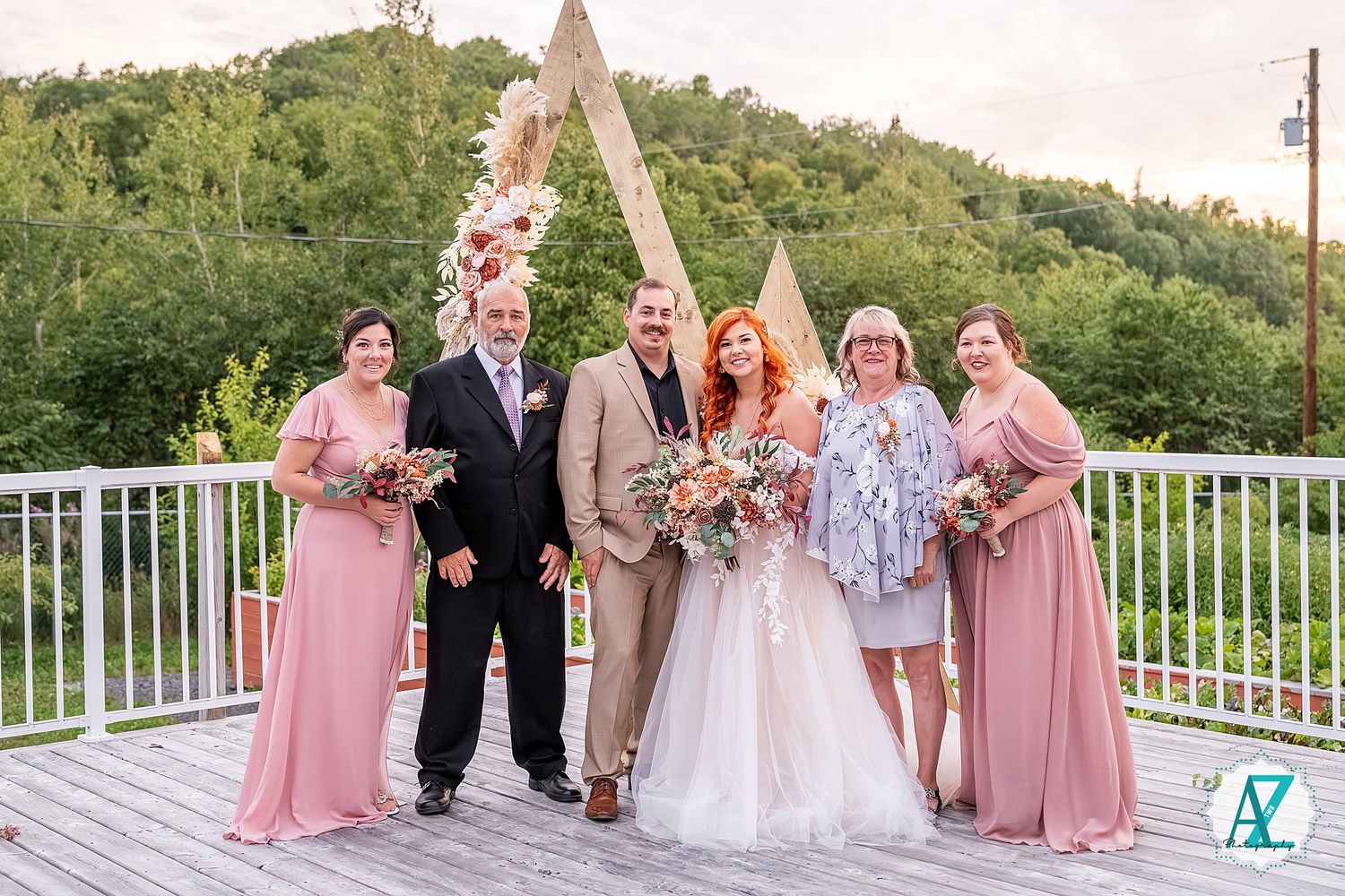 Wedding party members in pink and tan attire pose together on an outdoor deck at sunset.