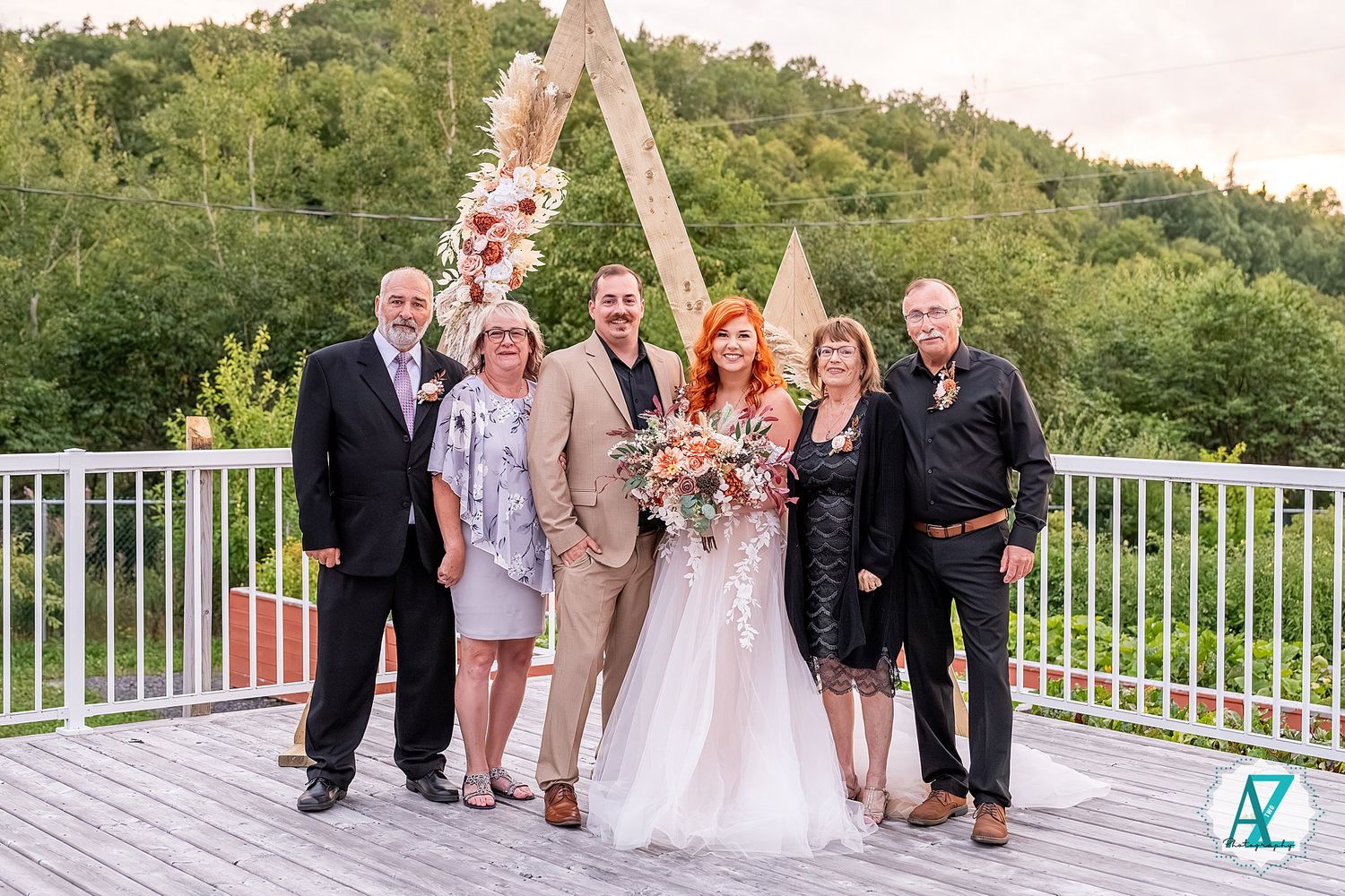 Wedding party gathers on outdoor deck with wooden triangle arch and mountain backdrop.