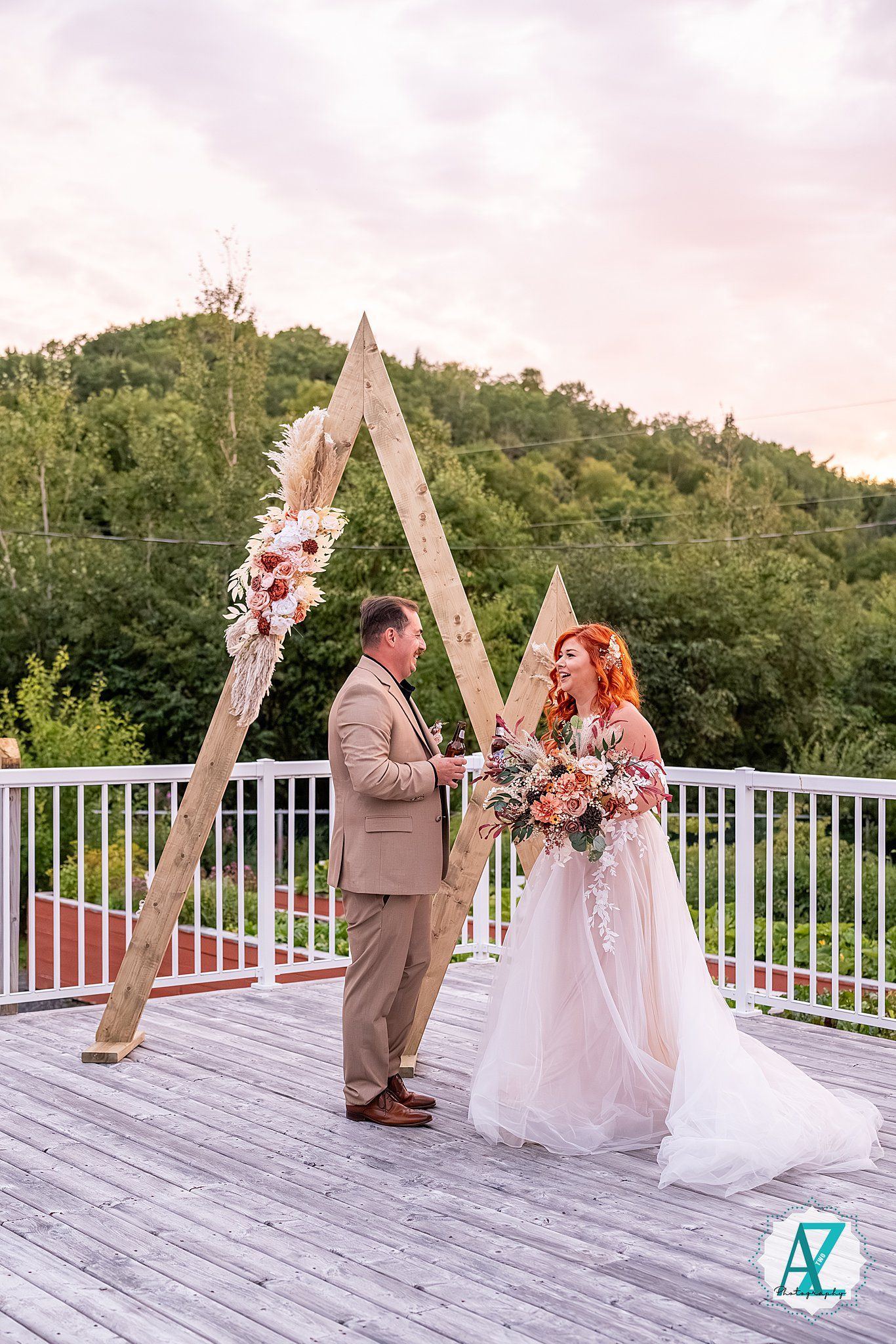 Wedding ceremony on white deck with triangular wooden arch decorated with pink and peach flowers against green hills.