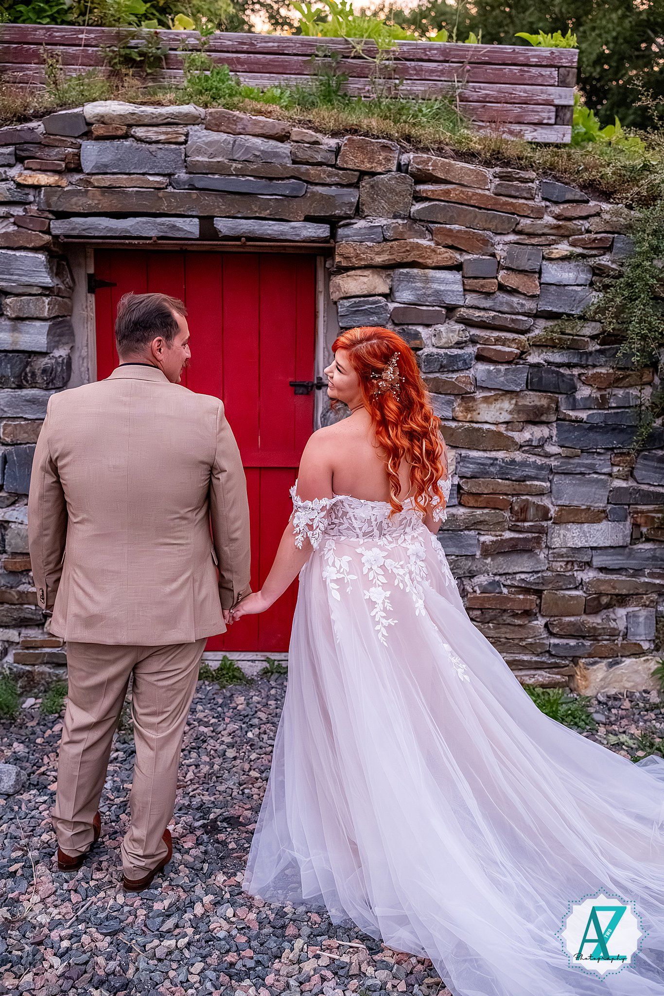A wedding couple walking together near a rustic stone wall with a red door.