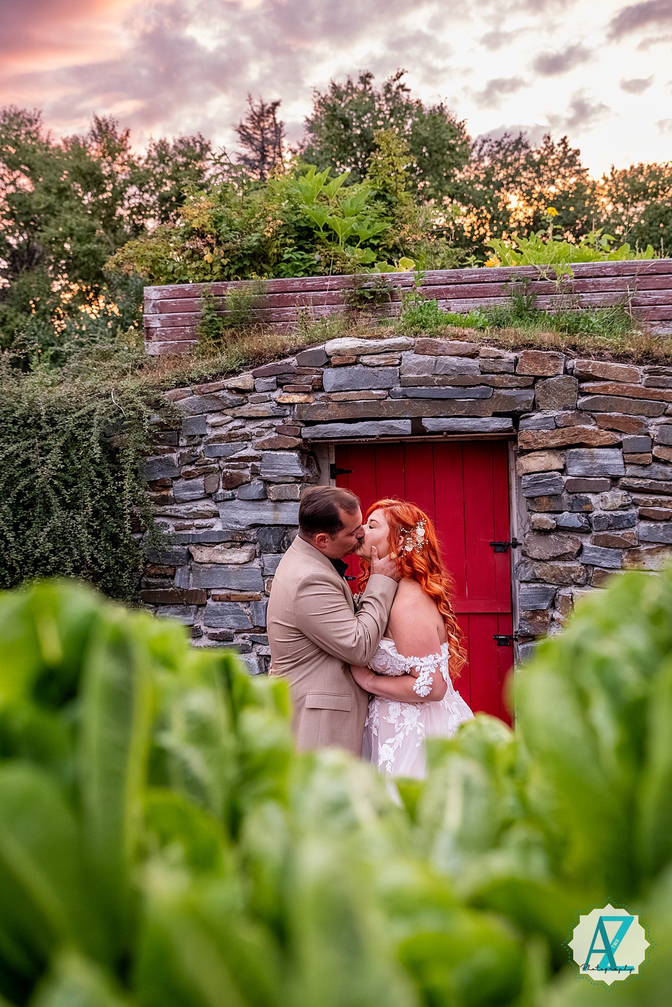 A romantic wedding couple embraces in front of a rustic stone wall with a red door during sunset.