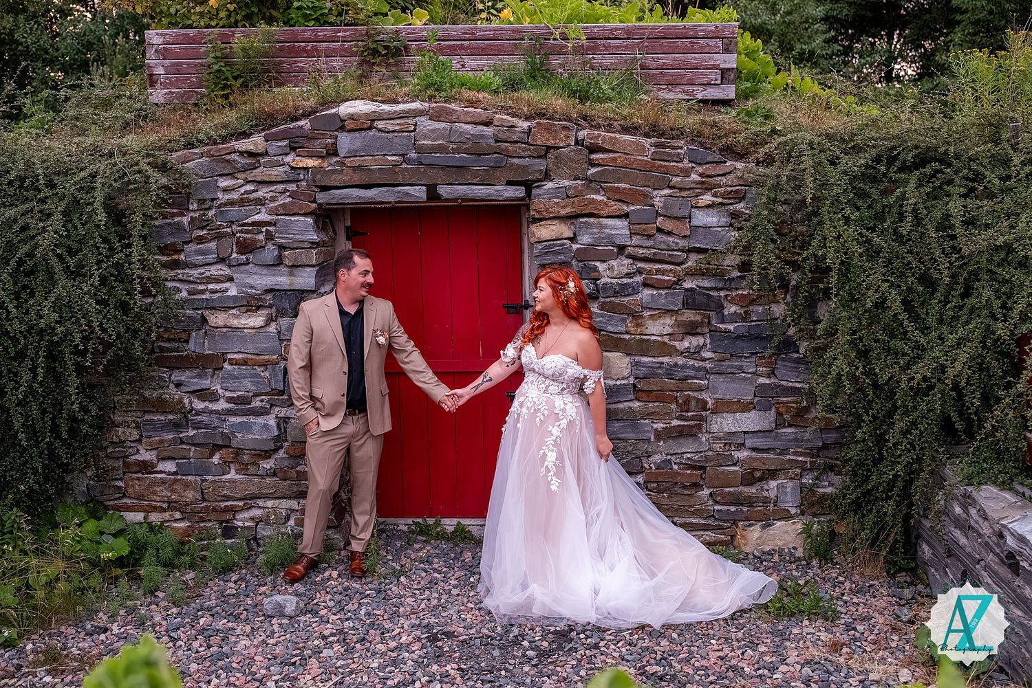 A newlywed couple poses in front of a rustic stone wall with a red door and greenery.