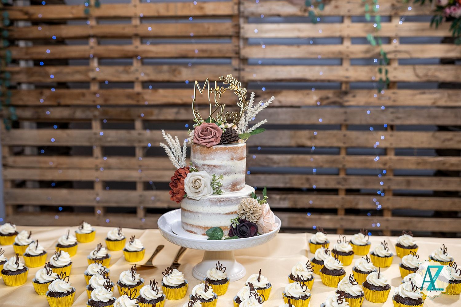 Wedding reception dessert table features cupcakes and cake tiers decorated in elegant style against wooden backdrop.