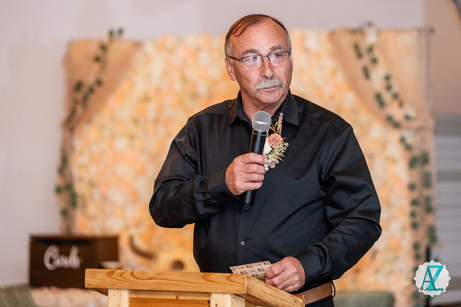 A speaker in a black button-down shirt gives a toast while holding a glass at an elegant wedding reception.