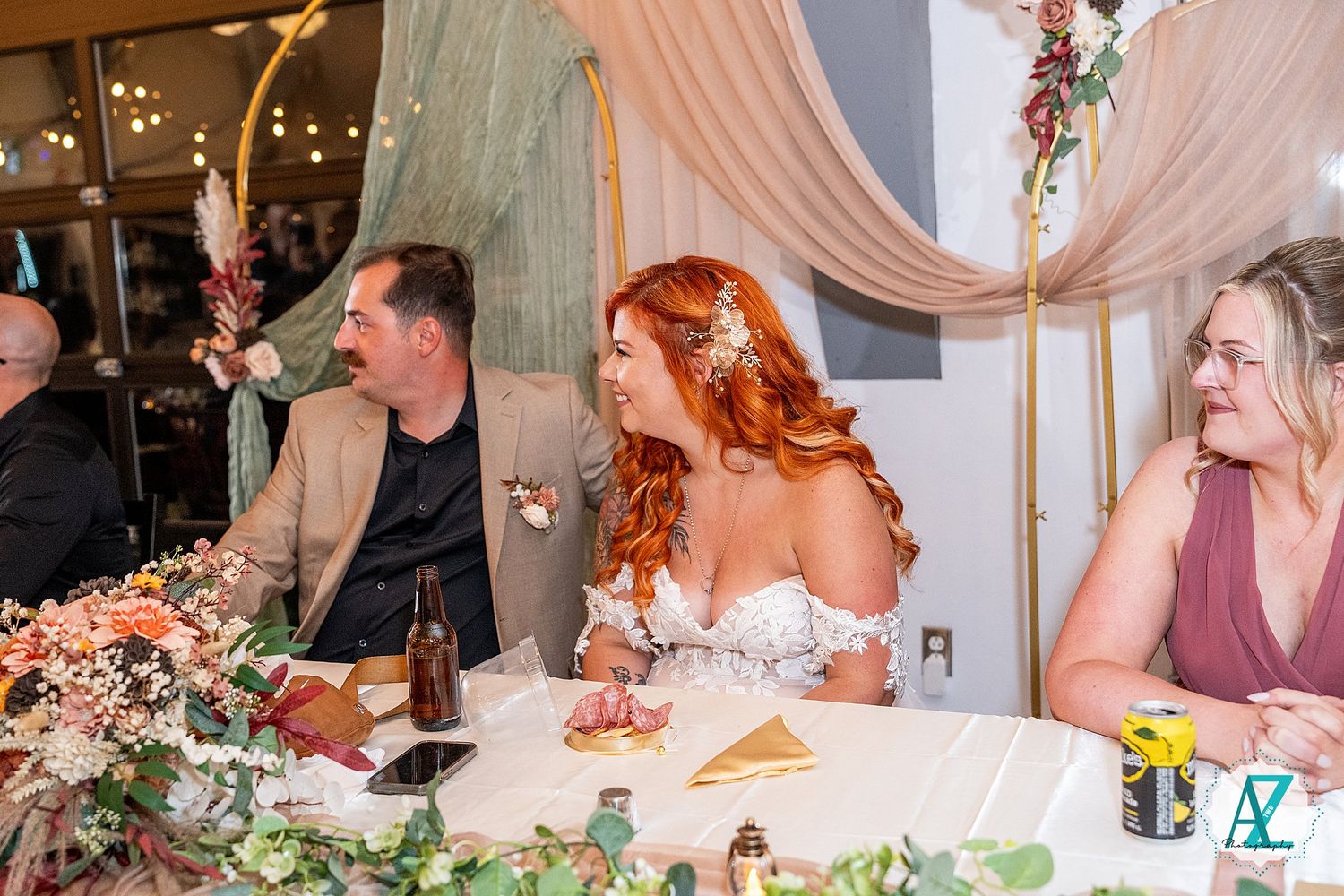 Wedding reception guests seated at a decorated dining table with floral arrangements and pink drapery.