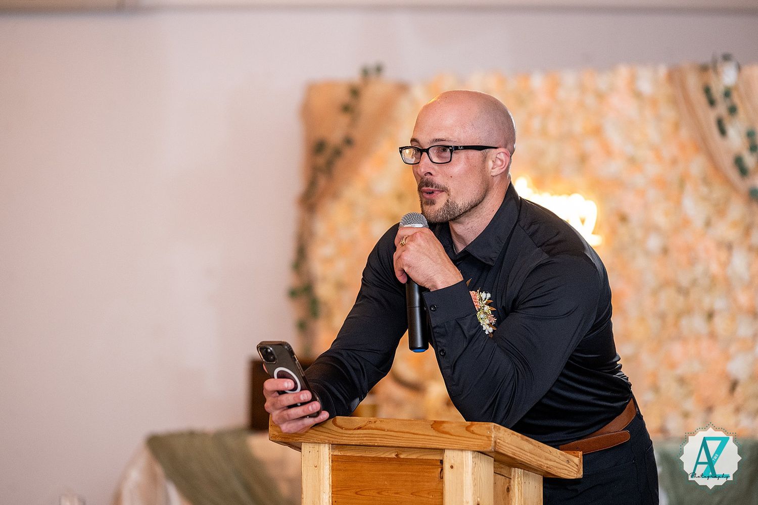 Dark-clad performer does a dramatic reading or poetry recital at a wooden podium in front of a patterned backdrop.