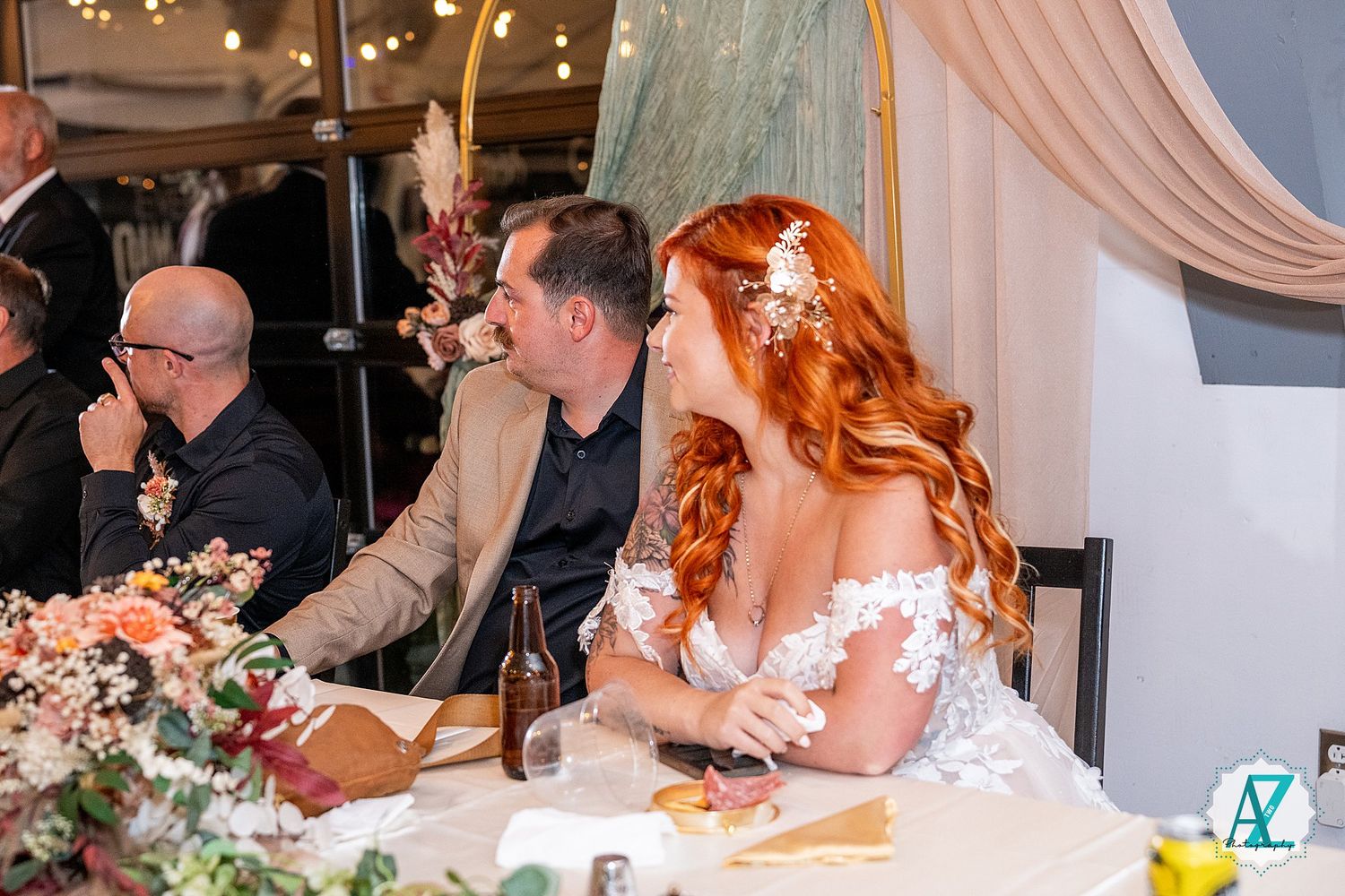 Wedding reception guests laugh and celebrate at decorated tables under warm lighting and floral arrangements.