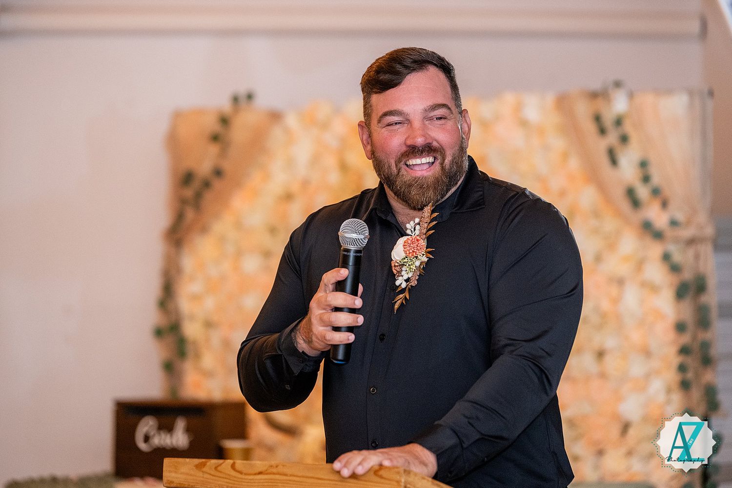 Wedding DJ in black attire performs and smiles while using microphone during reception celebration.