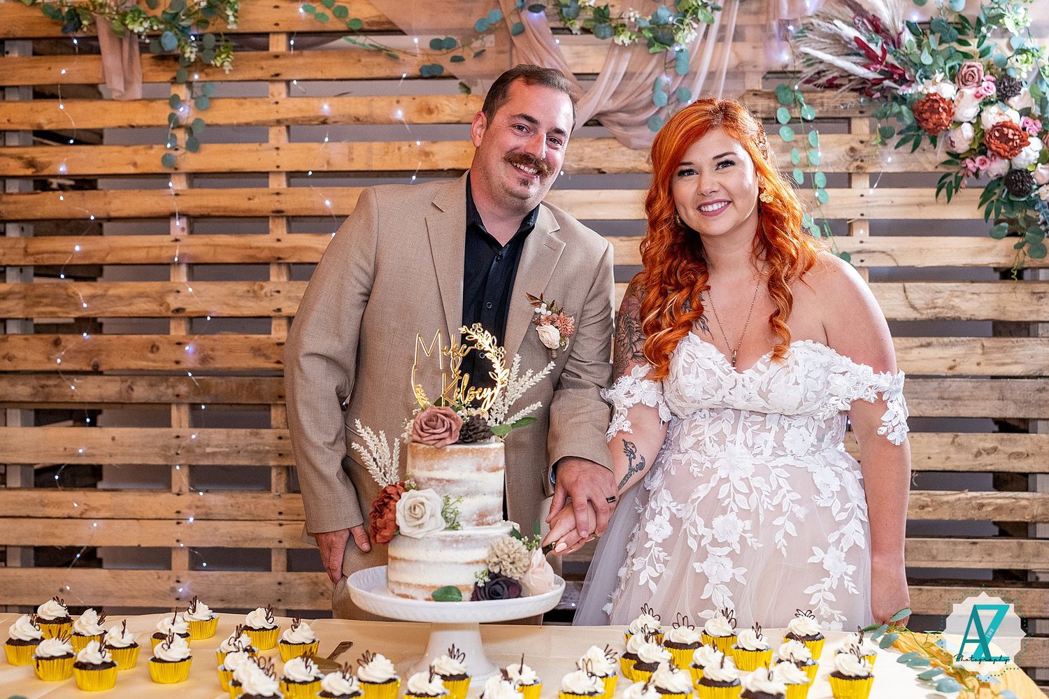 Newlyweds cut their wedding cake together at a rustic wood-paneled reception venue with string lights overhead.