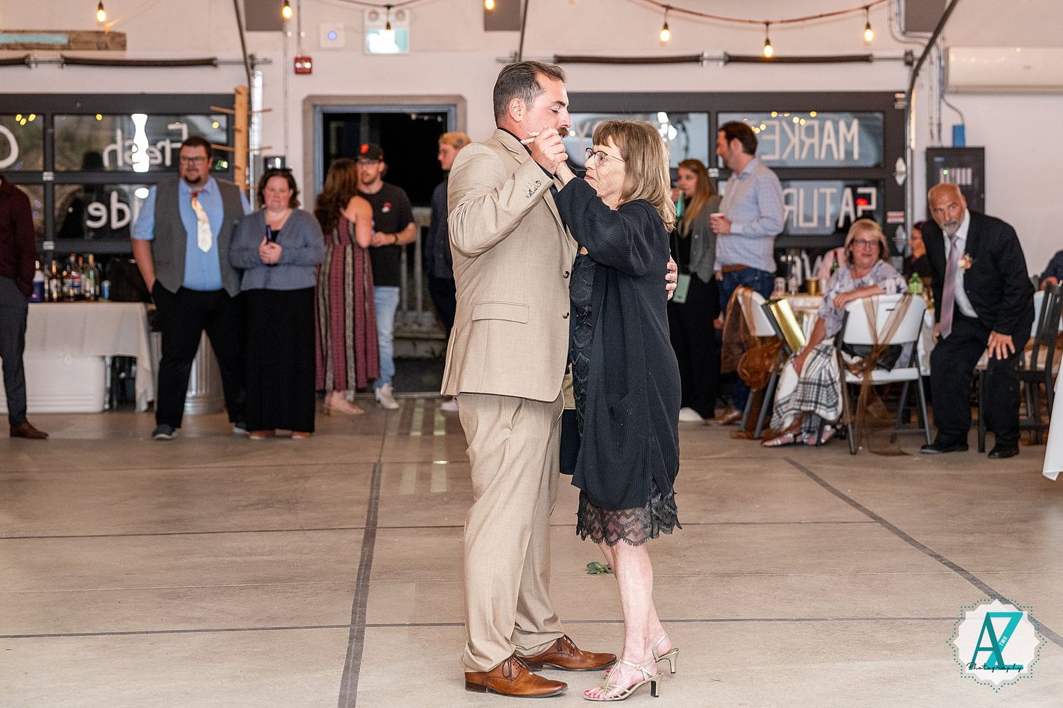 A couple dancing together at an indoor venue with string lights and other guests visible in the background.