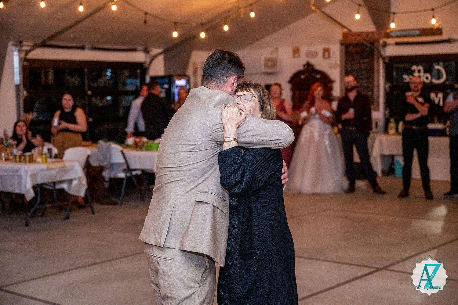 Wedding guests enjoy dancing in a rustic venue decorated with overhead string lights and white chairs.