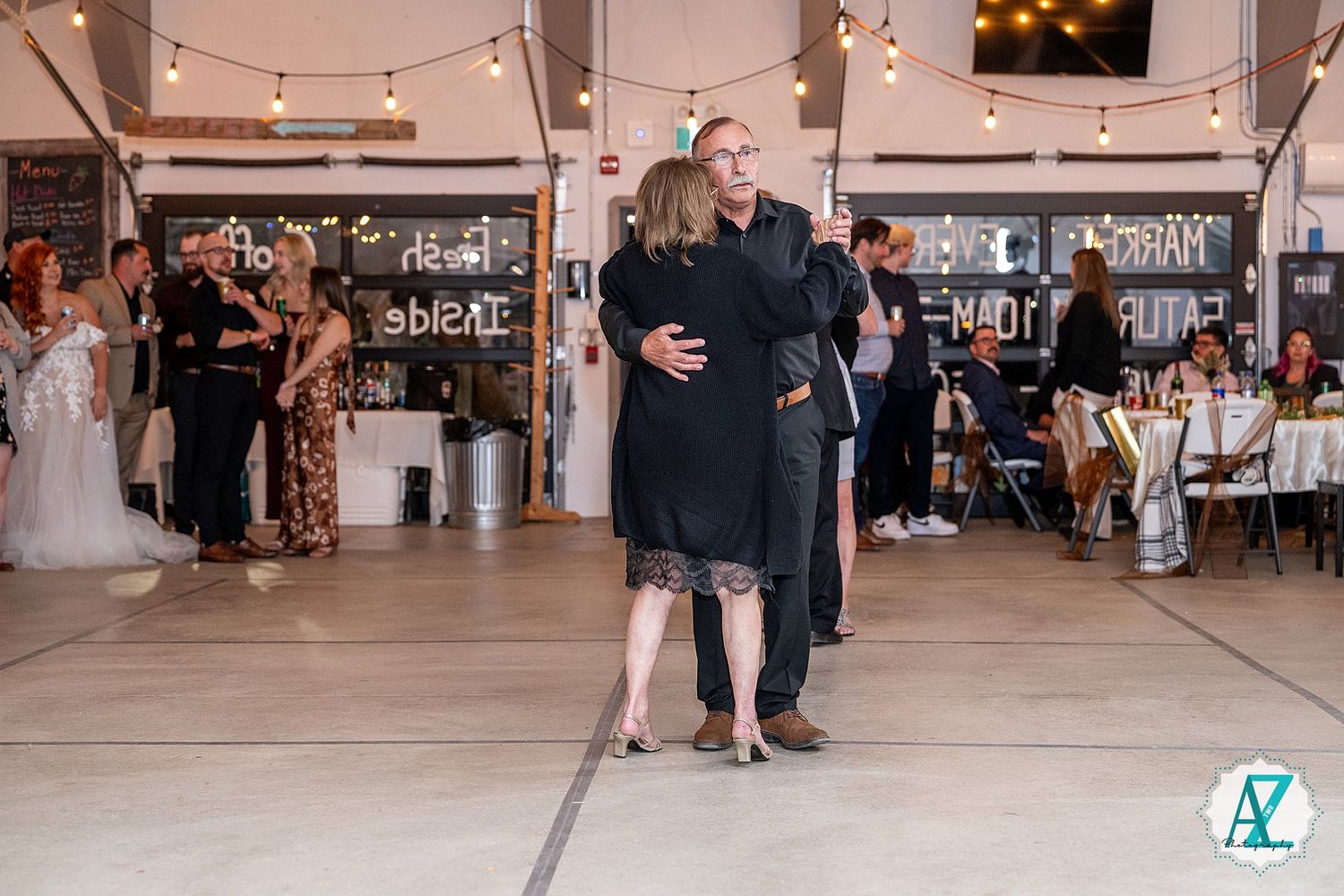 A couple shares an intimate dance moment under string lights at an indoor wedding reception venue.