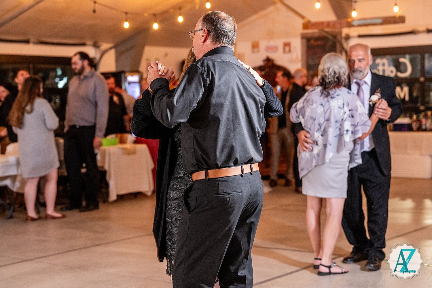 Indoor wedding venue with hanging lights and guests mingling on a wooden dance floor.