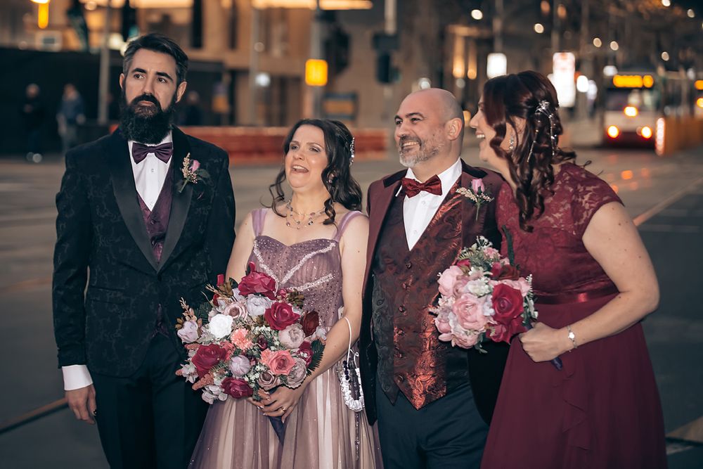 Wedding party in burgundy dresses and black tuxedos poses together with pink floral bouquets at night.