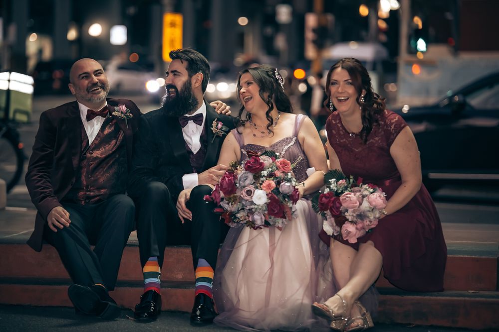 Wedding party members in formal attire gather outdoors at night holding floral bouquets.
