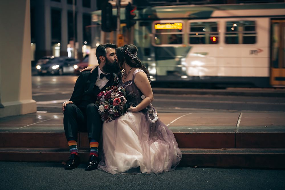 A couple in wedding attire sits together on benches at night with city lights in the background.
