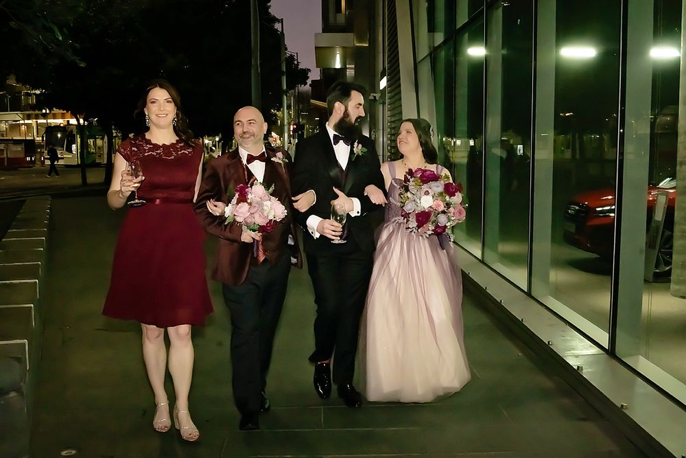 Wedding party members walk along illuminated glass windows at night in formal burgundy and black attire.