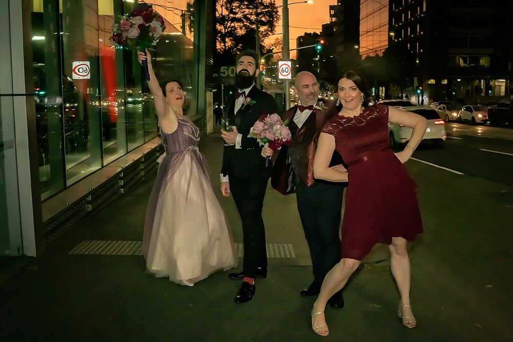 Wedding party members pose and laugh together outside a modern building at dusk.