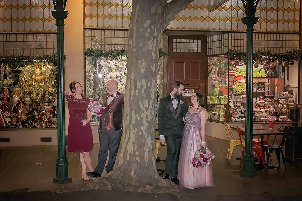 Multiple figures stand near tree trunks outside a building with warm lighting and vintage-style lampposts in the evening.