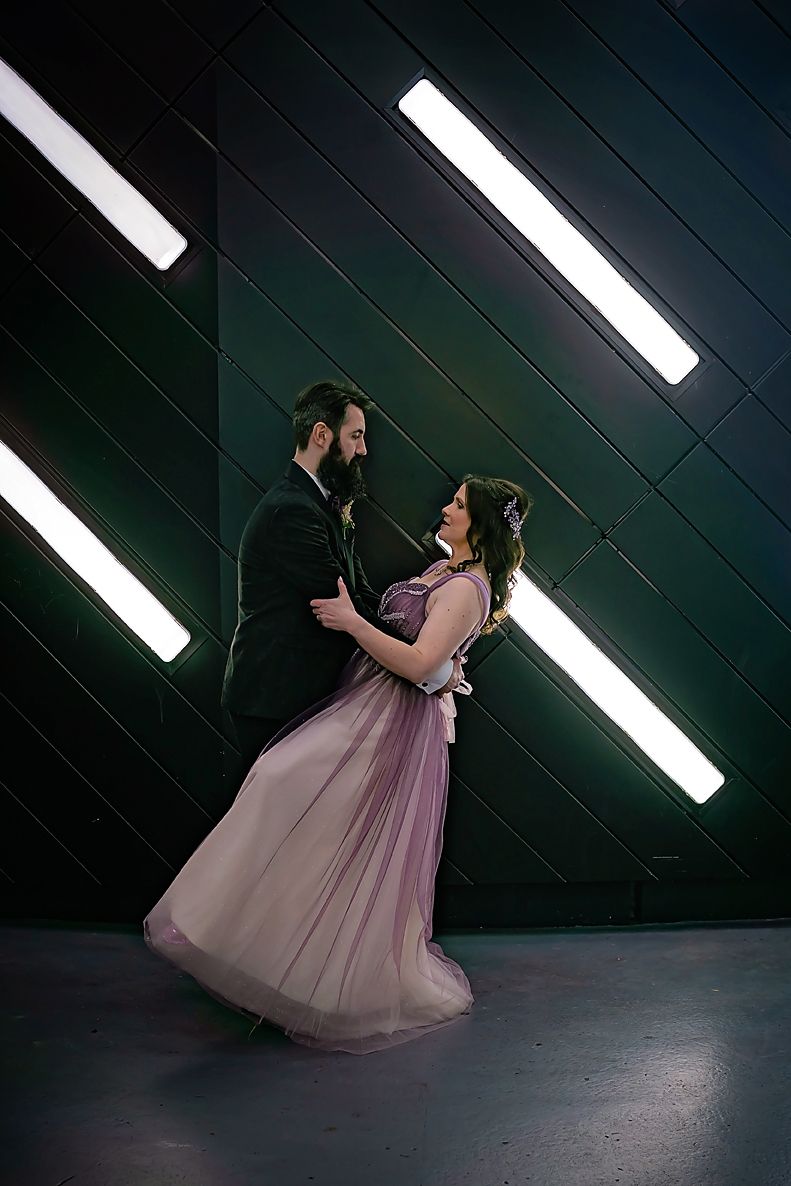 Couple dances dramatically in formal attire against dark wall with dramatic lighting.