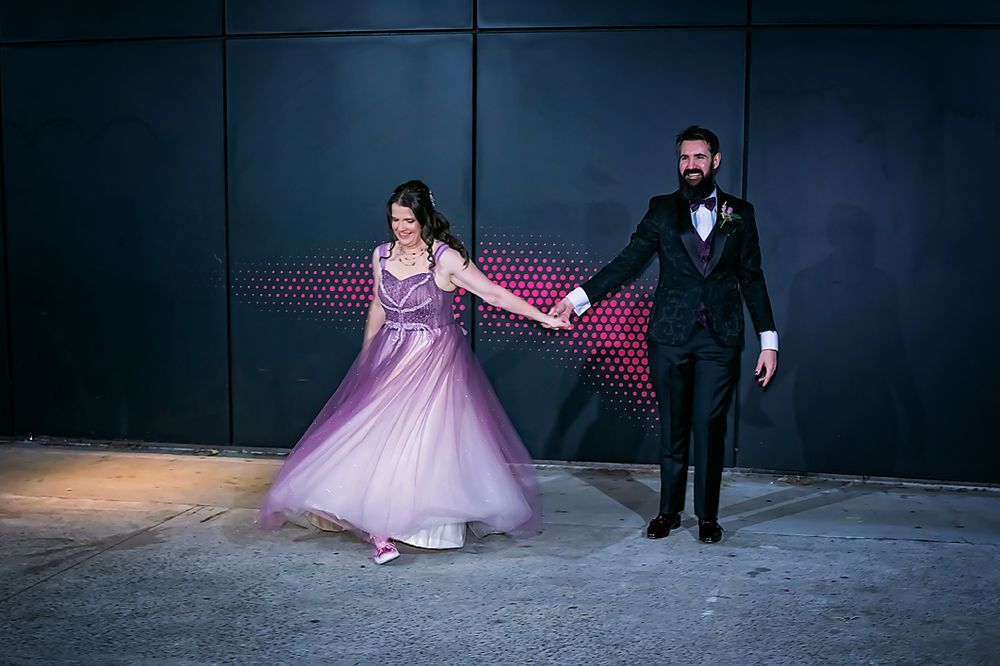 A couple in formal attire poses in a dark setting with purple accent lighting and dramatic shadows.