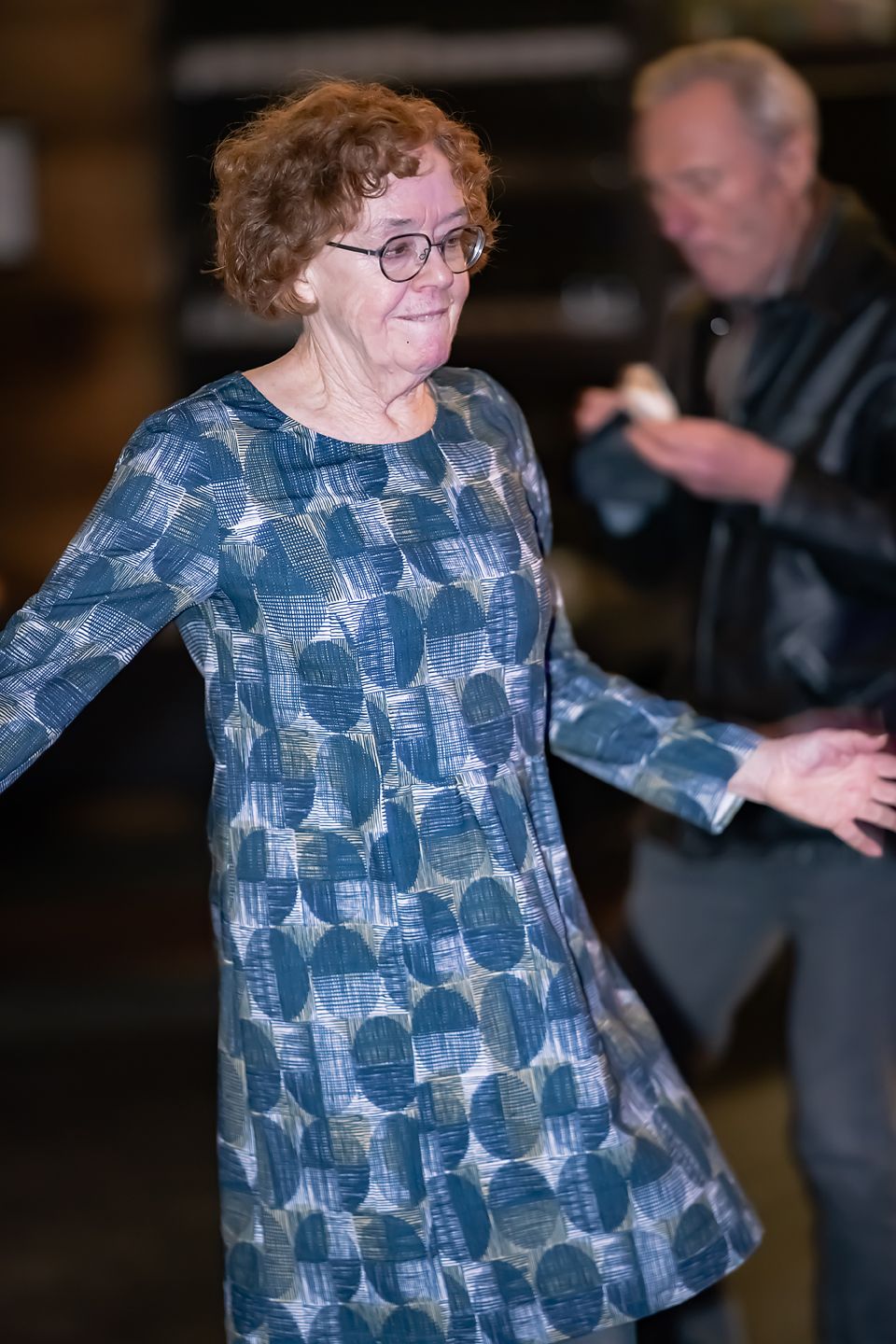 Person in a patterned blue and white blouse dances at an indoor event.