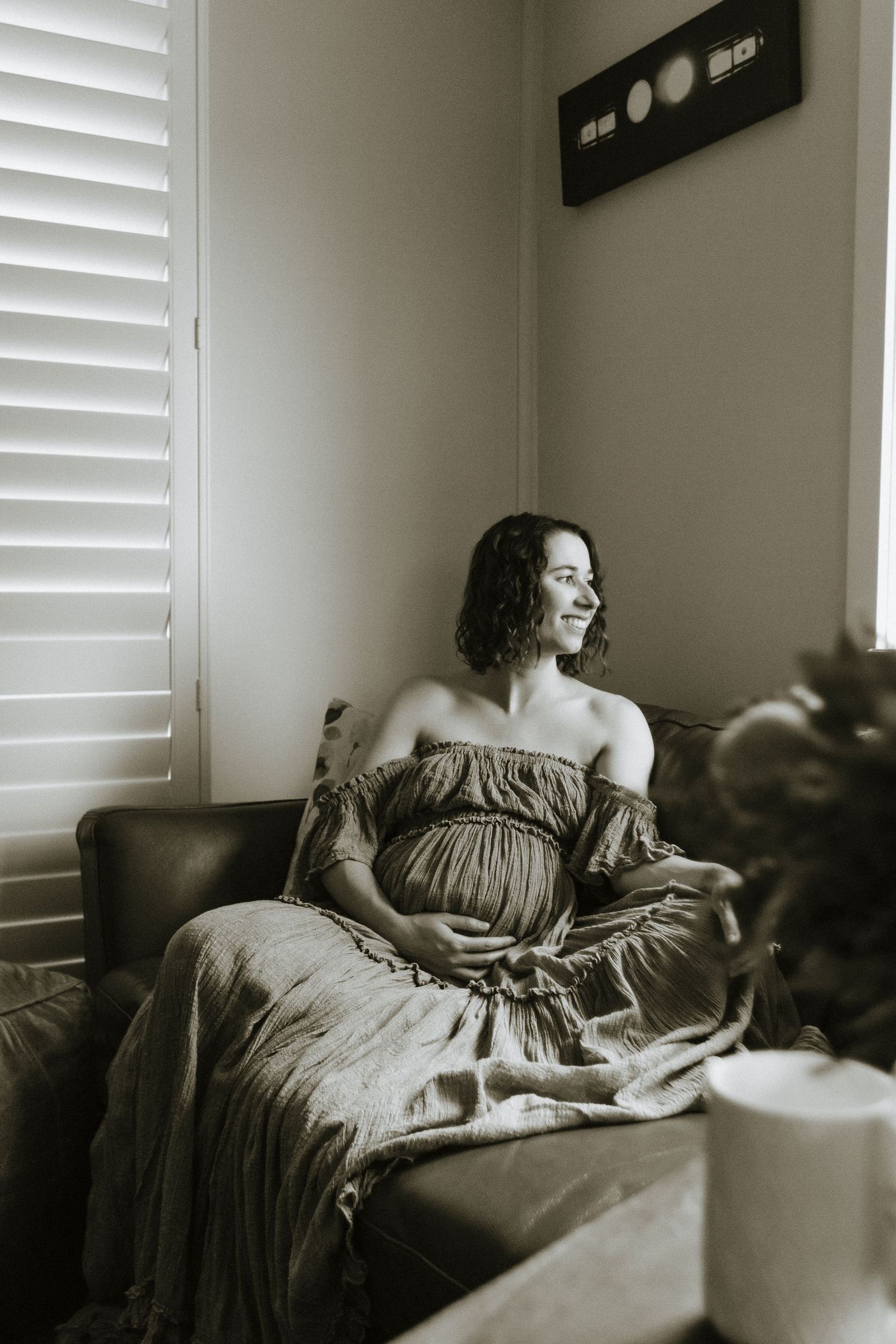Pregnant woman in an off-shoulder dress relaxes on couch by window with natural lighting.