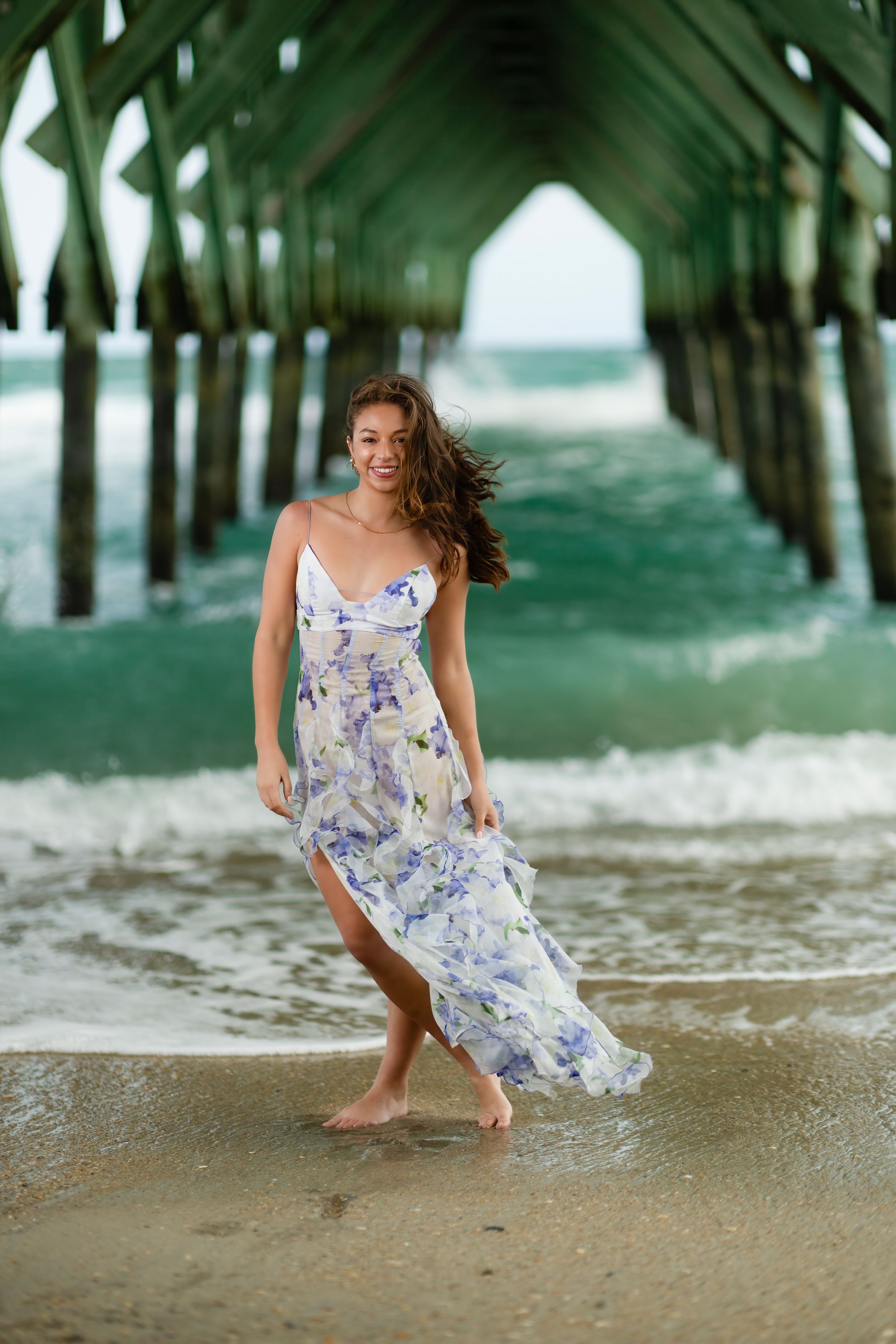 Long flowing floral dress catches the breeze under a pier as waves crash on the beach.