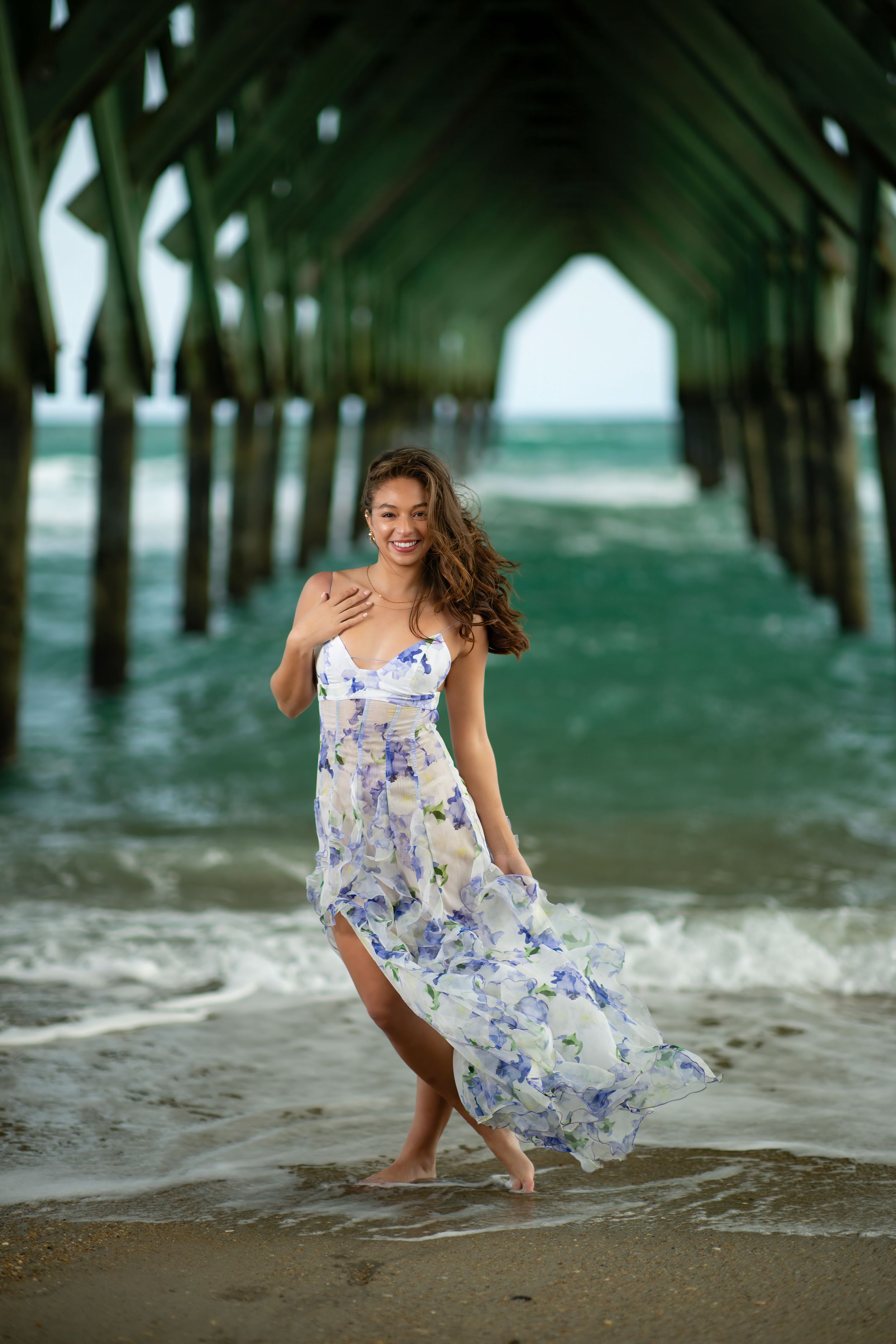 A flowing floral dress catches the ocean breeze under a pier as waves roll in on a summer beach day.