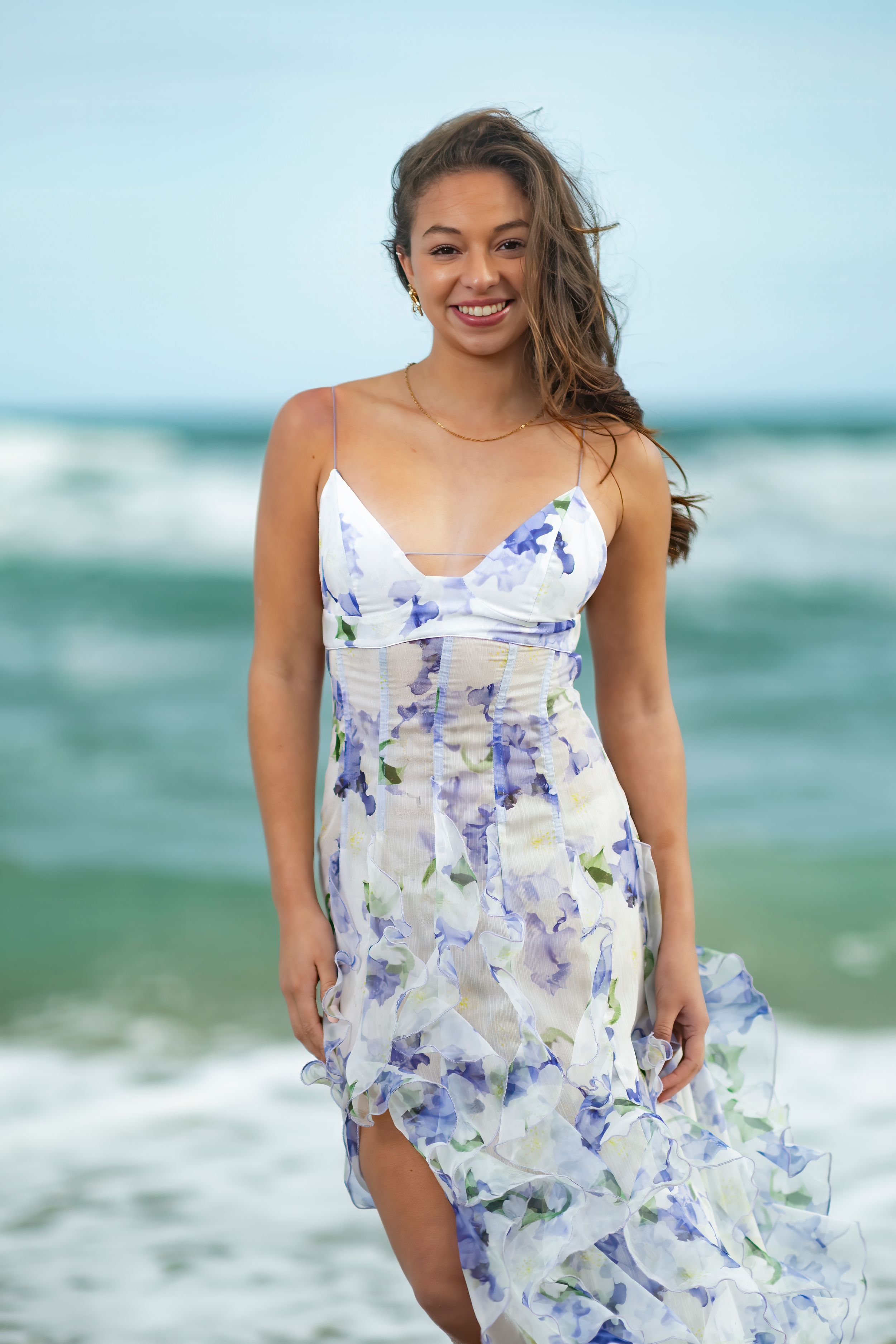 A woman in a flowing white and blue floral dress poses on a beach with turquoise waves in the background.