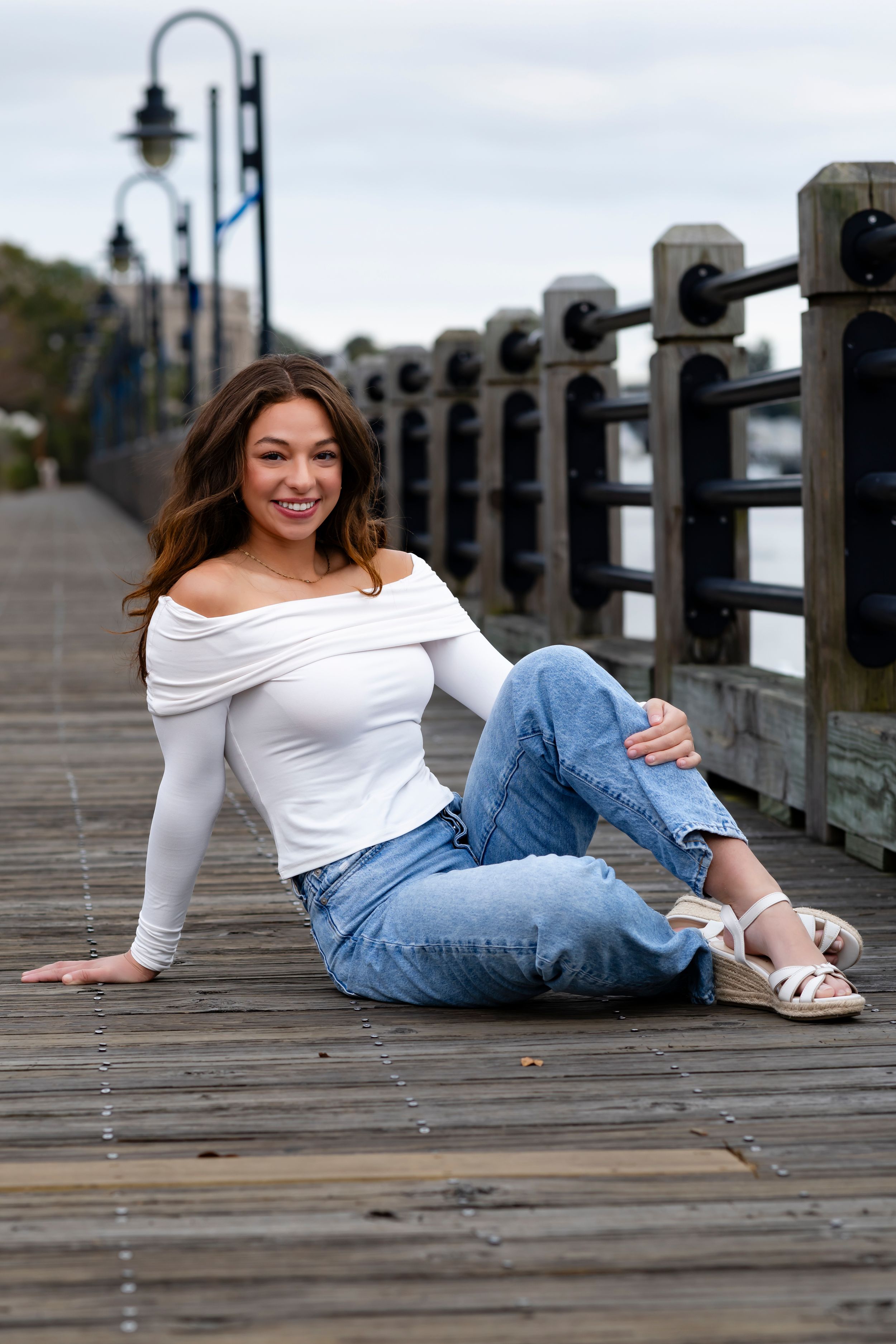 Casual portrait on wooden boardwalk with white off-shoulder top, jeans and sandals against waterfront railing and lamp posts.