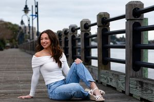 Person in white sweater and jeans sitting casually on wooden pier railing on cloudy day.